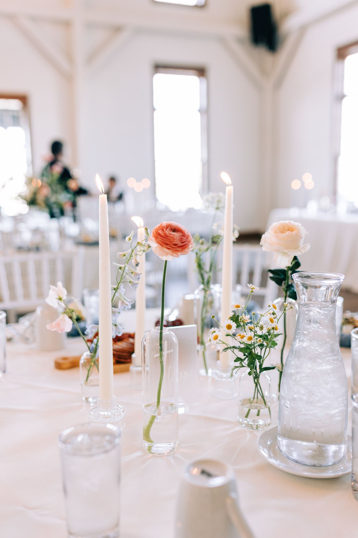 Minimalist wedding table setting with tall taper candles and single stem flowers in glass bud vases.