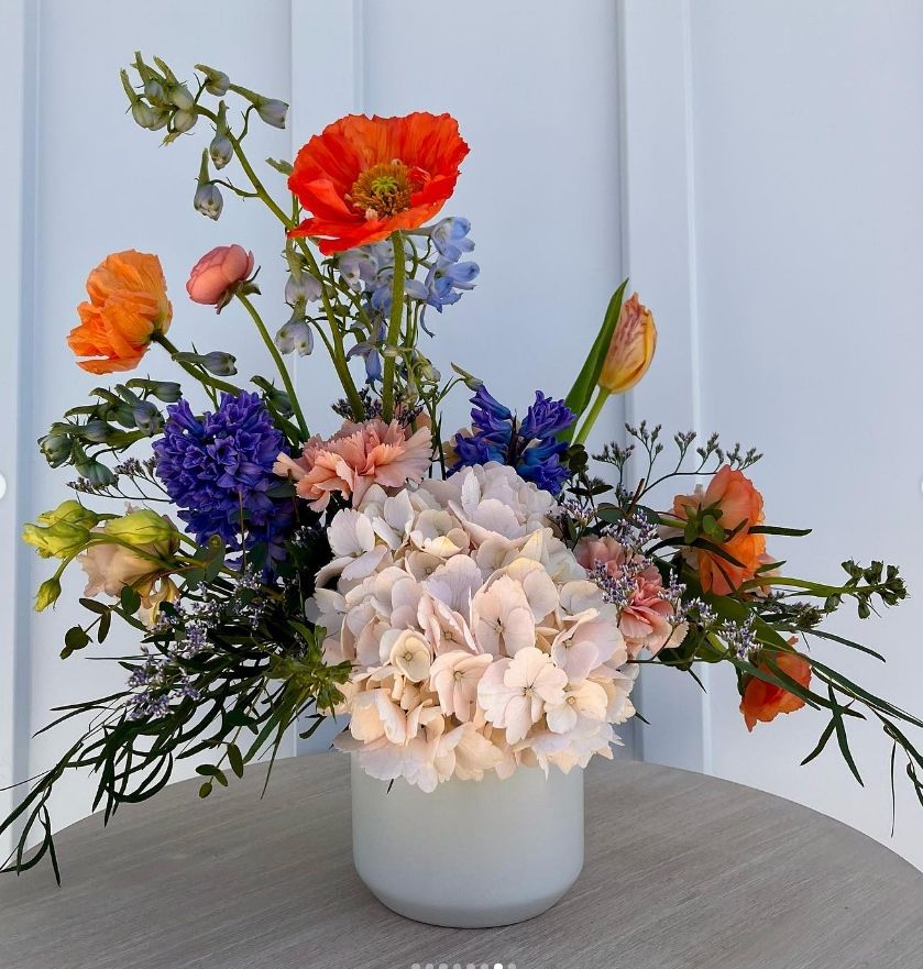 Cheerful arrangement with orange poppies, blue delphiniums, and pink hydrangea in white vase.