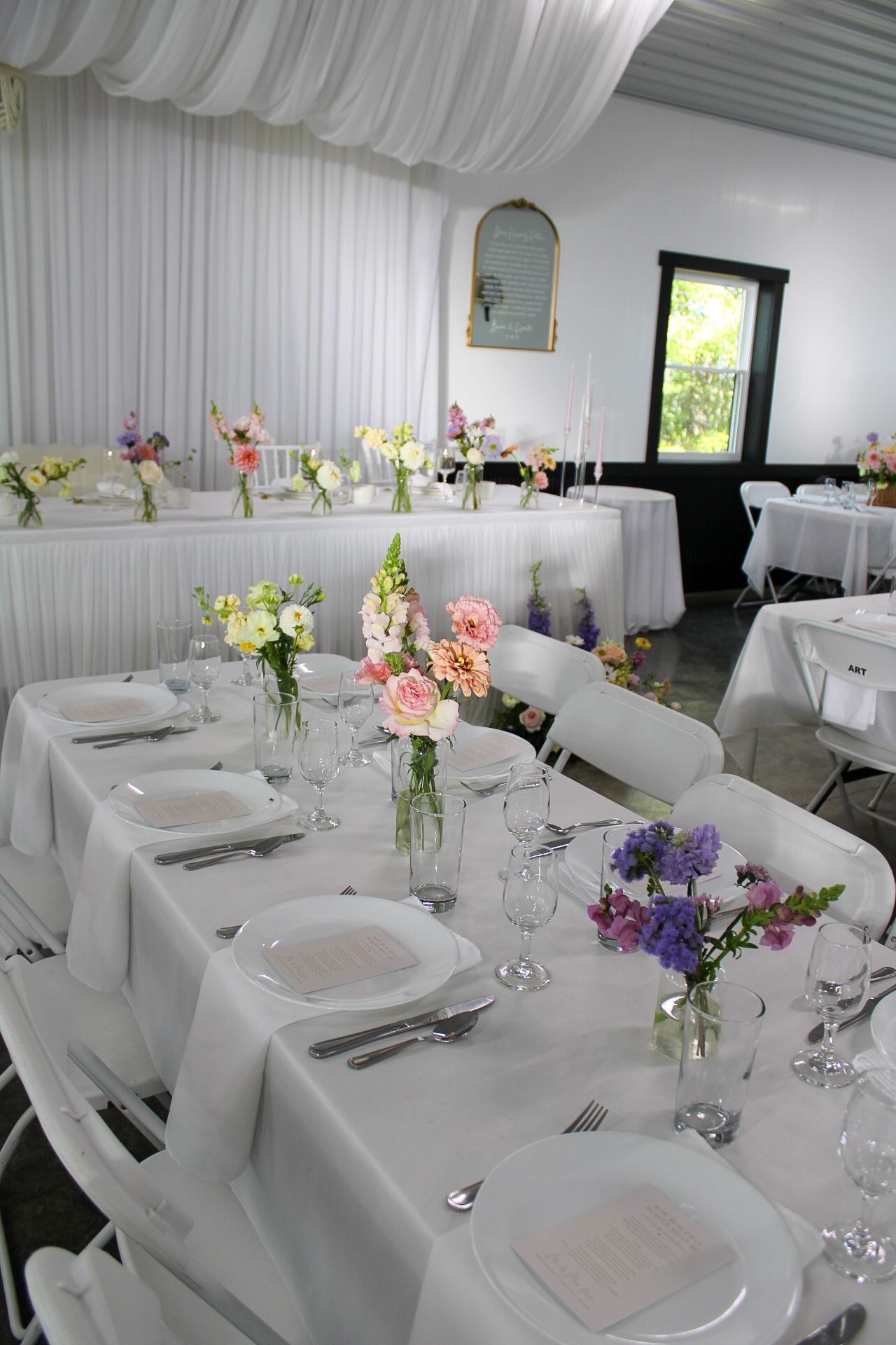 Elegant white wedding reception tables with floral centerpieces featuring pink, purple and white flowers in glass vases.