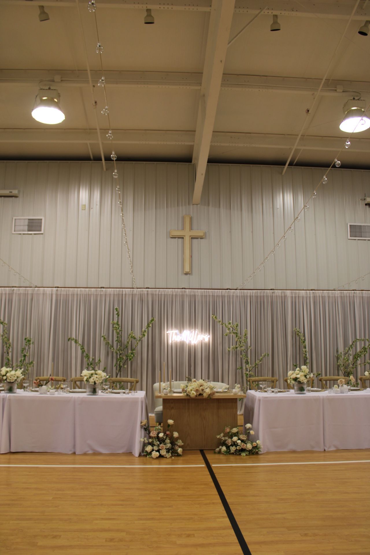 Church gymnasium setup for wedding reception with white tablecloths and wooden cross on the wall.