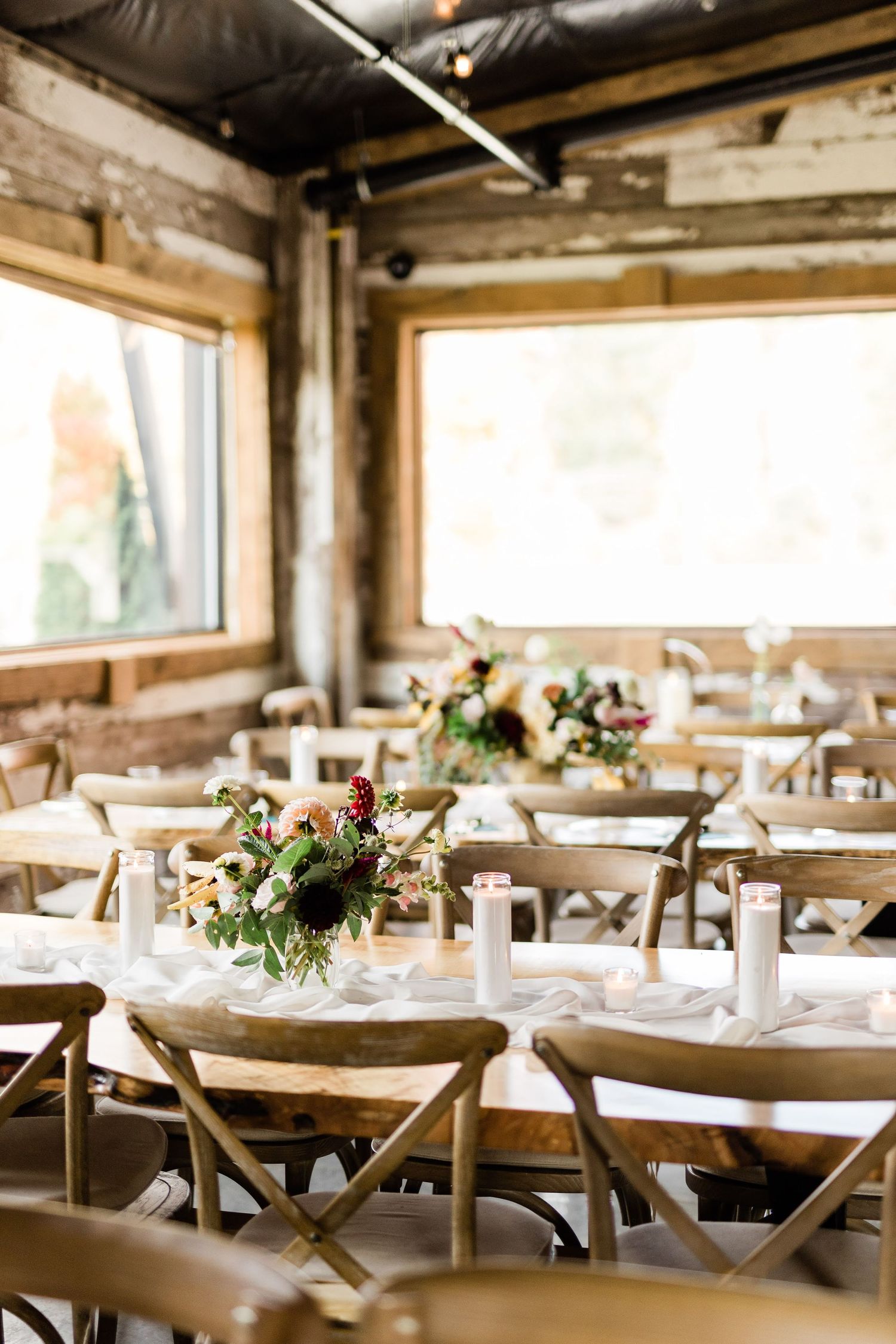 Rustic wedding reception setup with crossback chairs and floral centerpieces in sunlit venue.