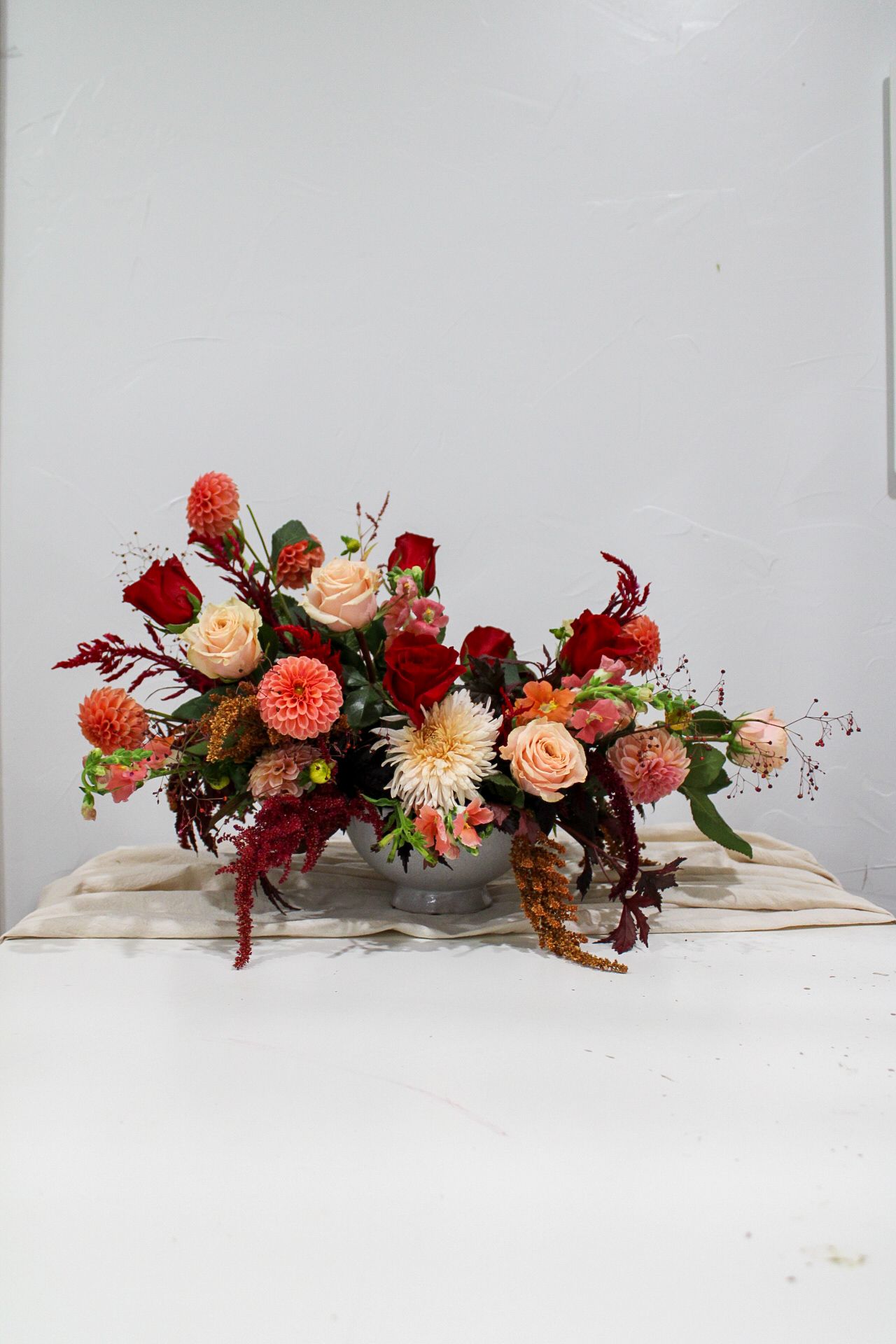 Dramatic floral centerpiece with red and peach roses, dahlias and trailing foliage on white surface.