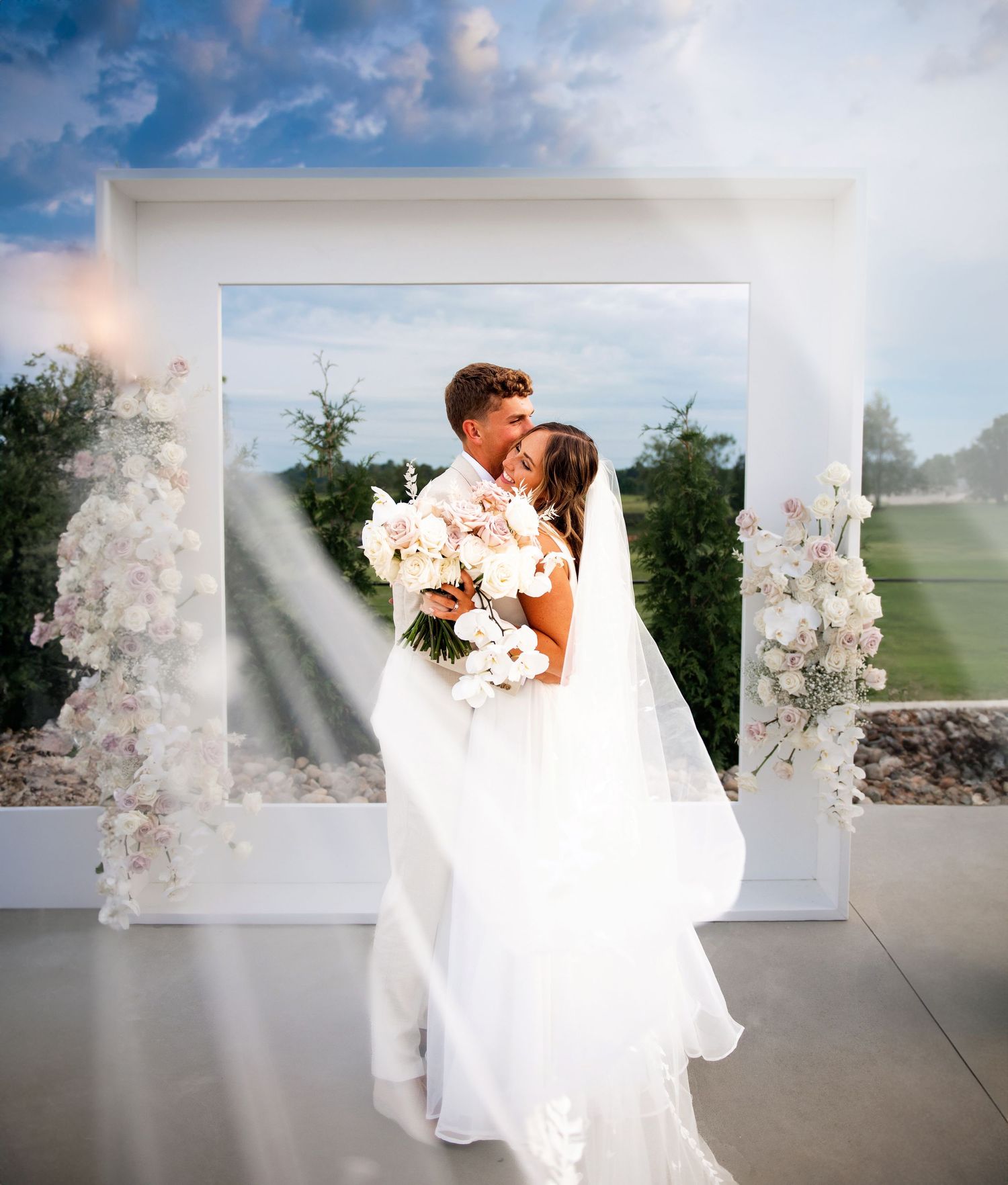 A romantic outdoor wedding ceremony featuring a white floral arch and flowing bridal gown in natural light.