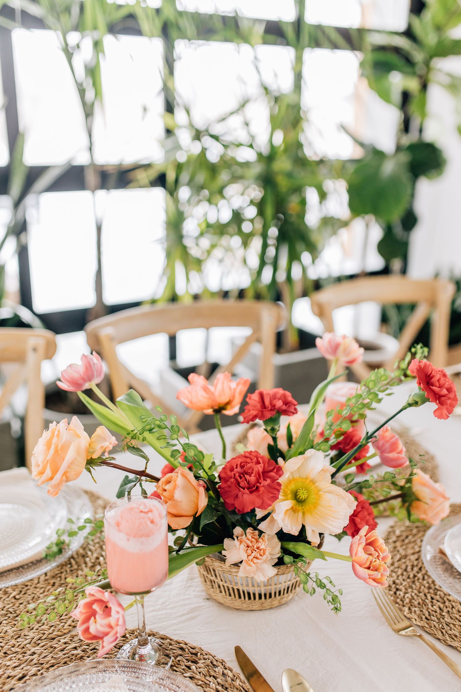 Bright coral and pink floral arrangement on table with woven placemats and elegant place settings.