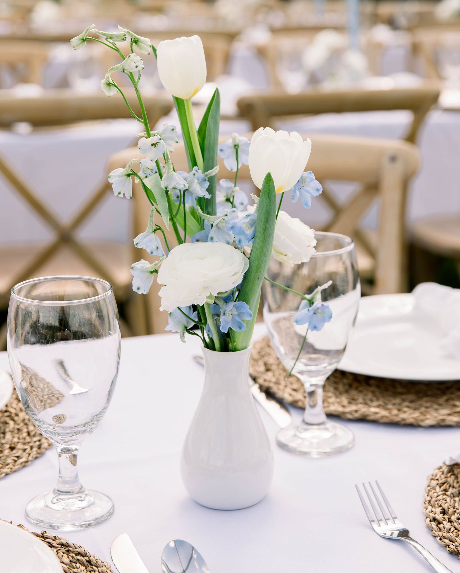 Elegant white tulips and blue delphinium flowers in a small white vase on a formal dining table with place settings.