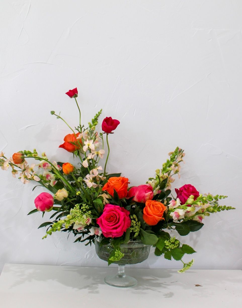 Vibrant arrangement of orange and pink roses with greenery in a glass pedestal bowl.