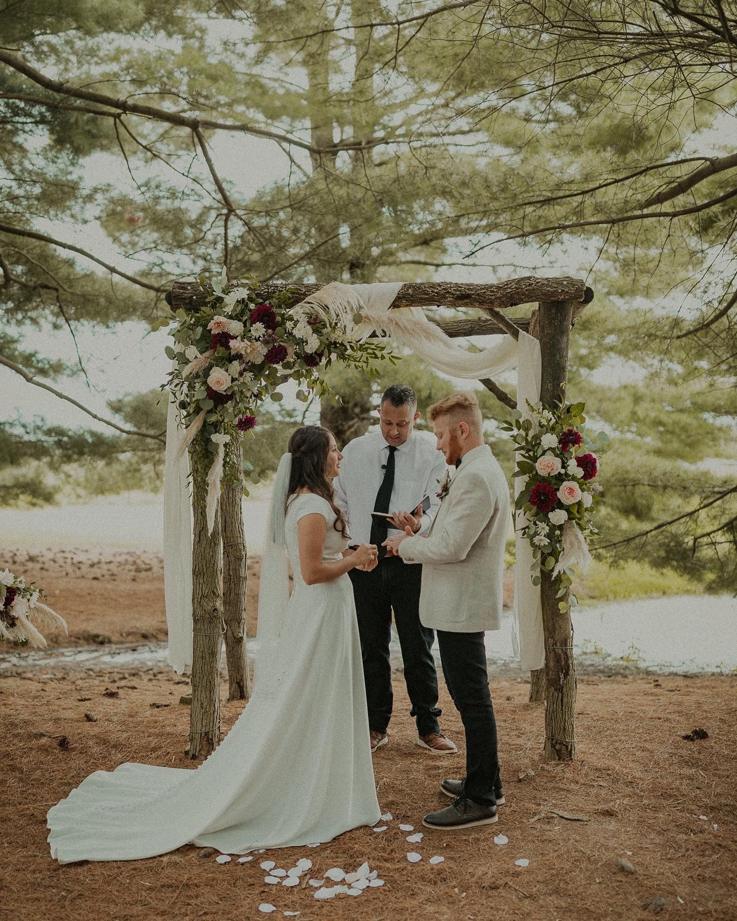 Rustic wedding ceremony under wooden arch with pink and burgundy flowers in forest setting.