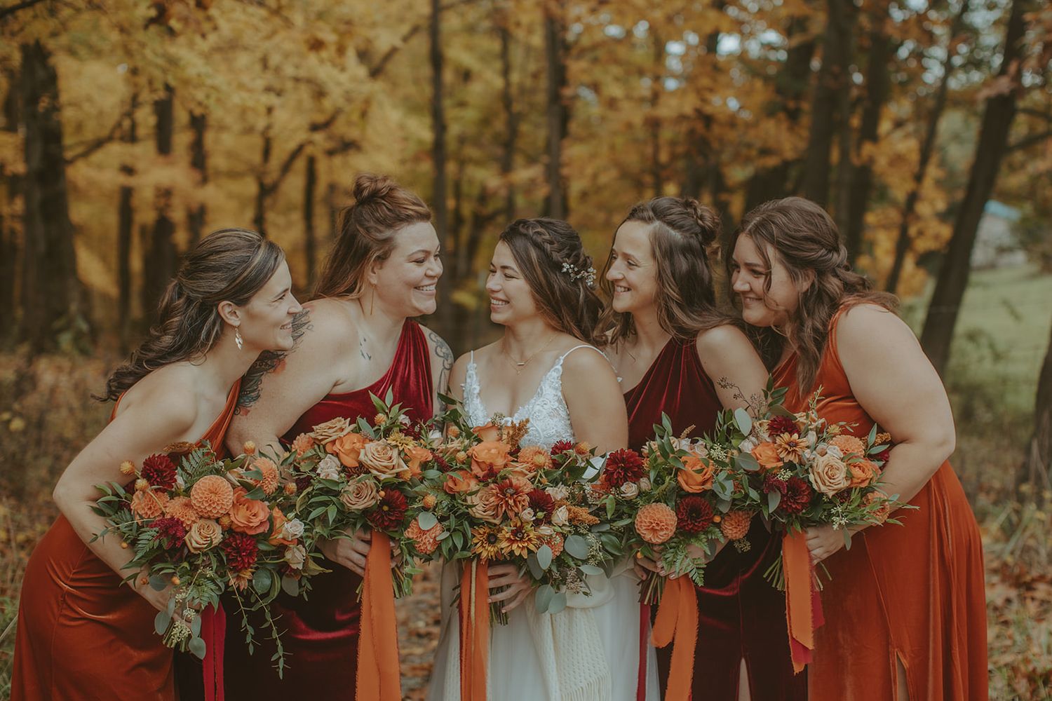 Wedding party in burgundy dresses huddles close together holding autumn-colored bouquets.