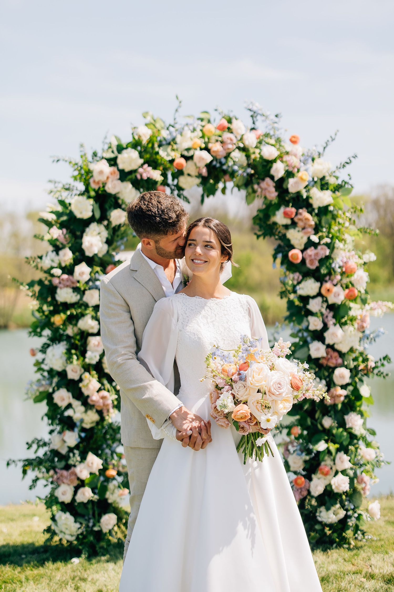Romantic outdoor wedding ceremony with floral arch and lakeside setting in soft pastel colors.