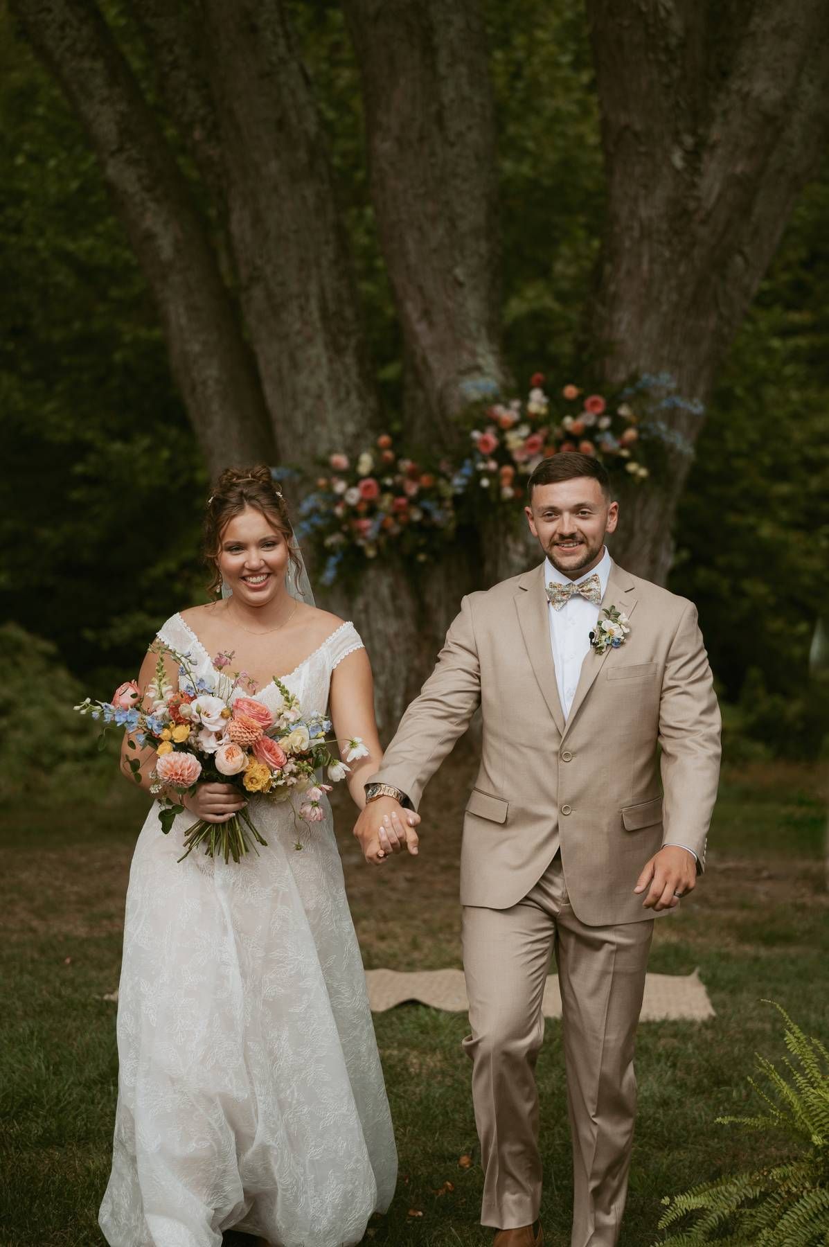 A couple shares joyful candid moments in a wooded outdoor setting wearing wedding attire in tan and white.