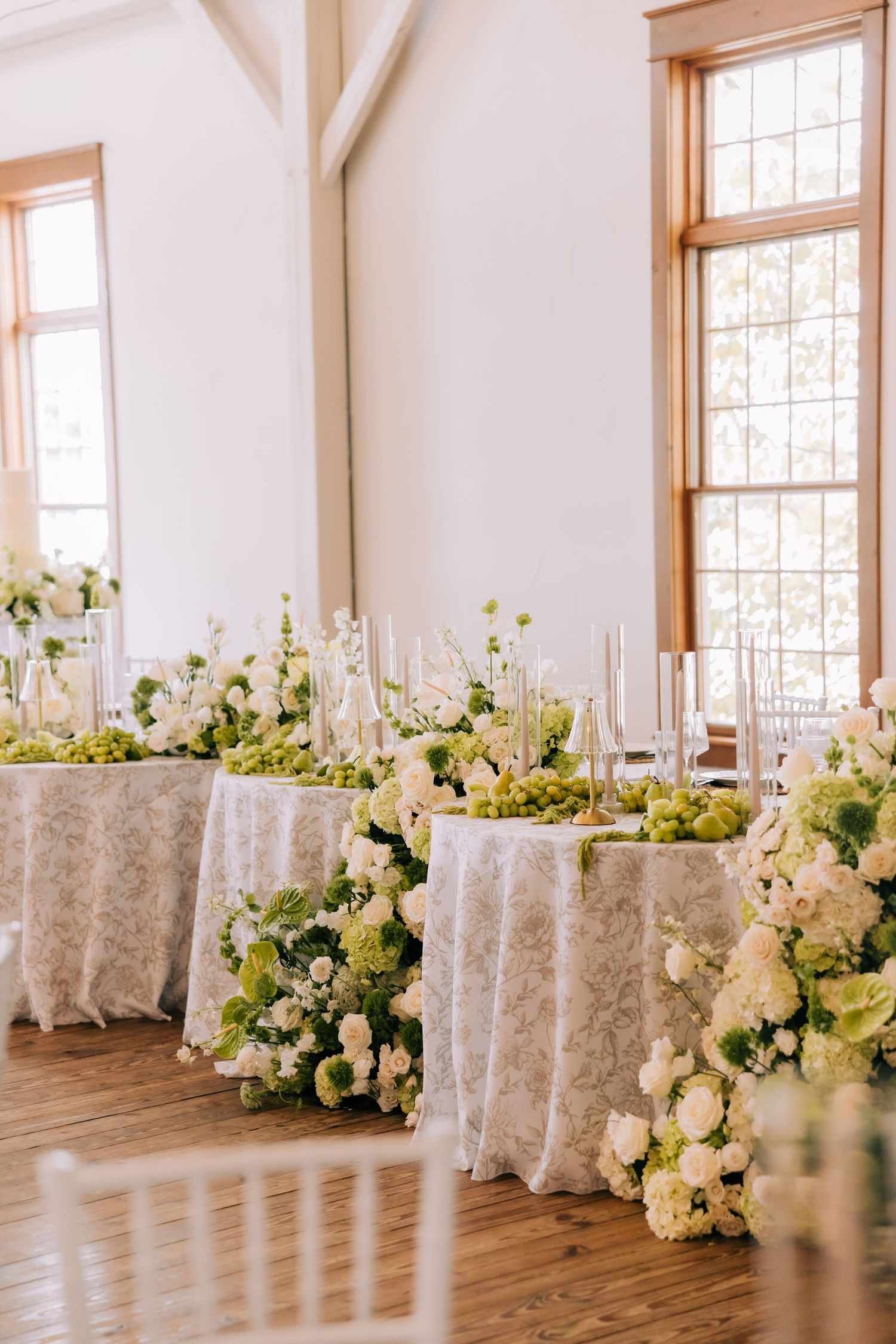 Long banquet tables with elegant white tablecloths and lush floral centerpieces in a bright sunlit reception venue.
