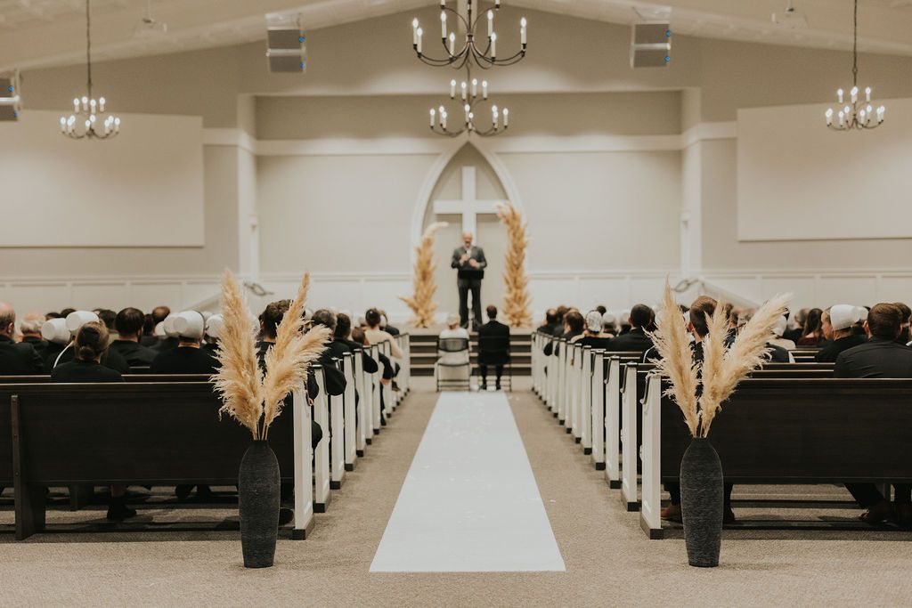A modern white wedding chapel interior with crystal chandeliers and wooden pews lining both sides of the aisle.