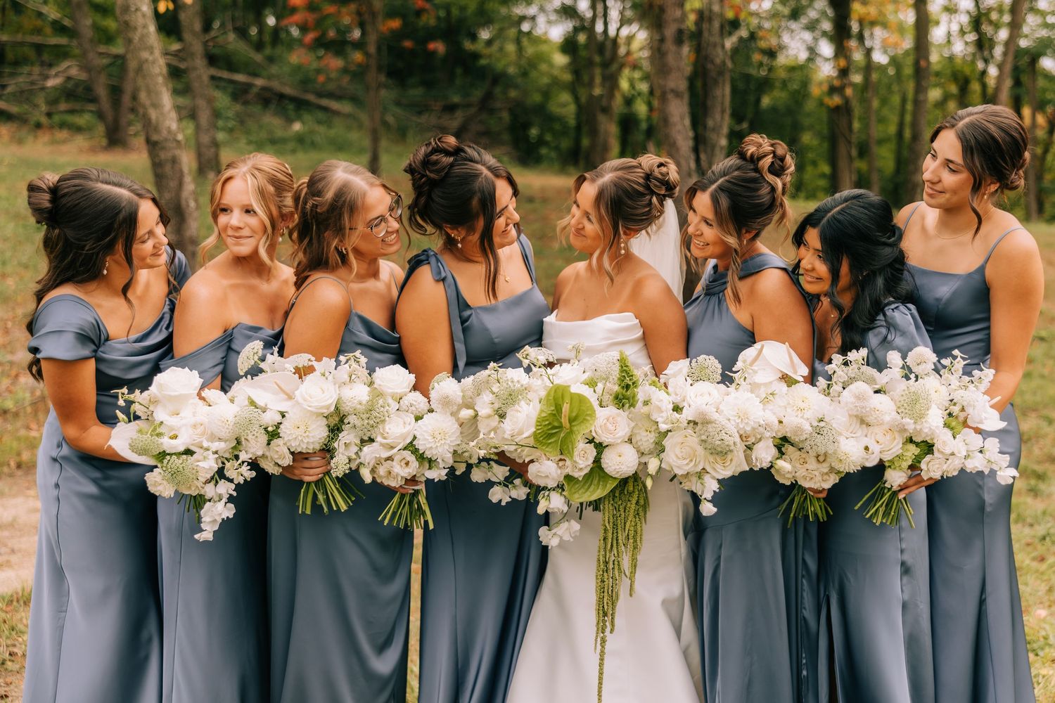 Bridesmaids in dusty blue dresses hold elegant white floral bouquets while standing together outdoors.
