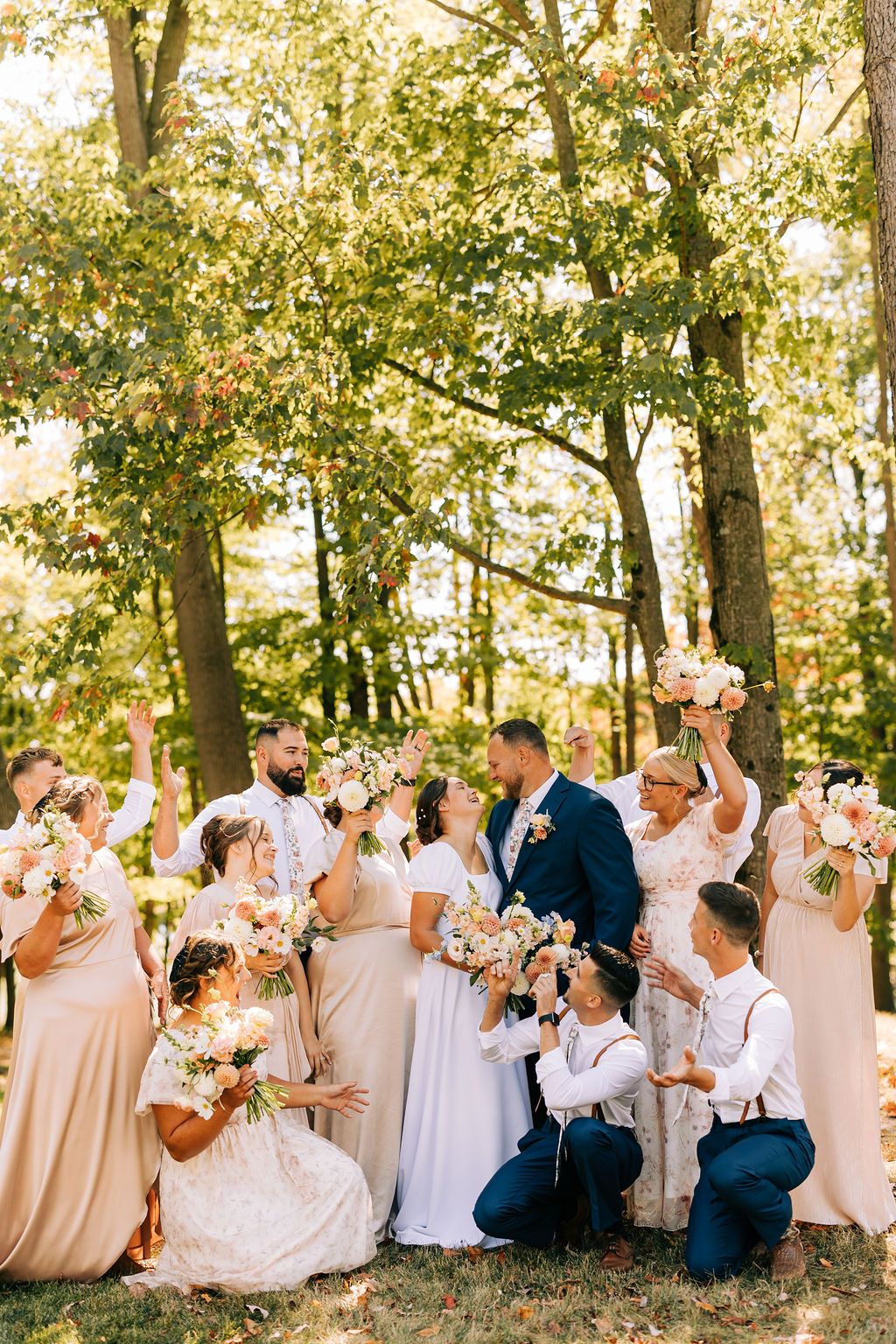 Wedding party in light beige and navy attire poses together in a wooded outdoor setting with natural sunlight.