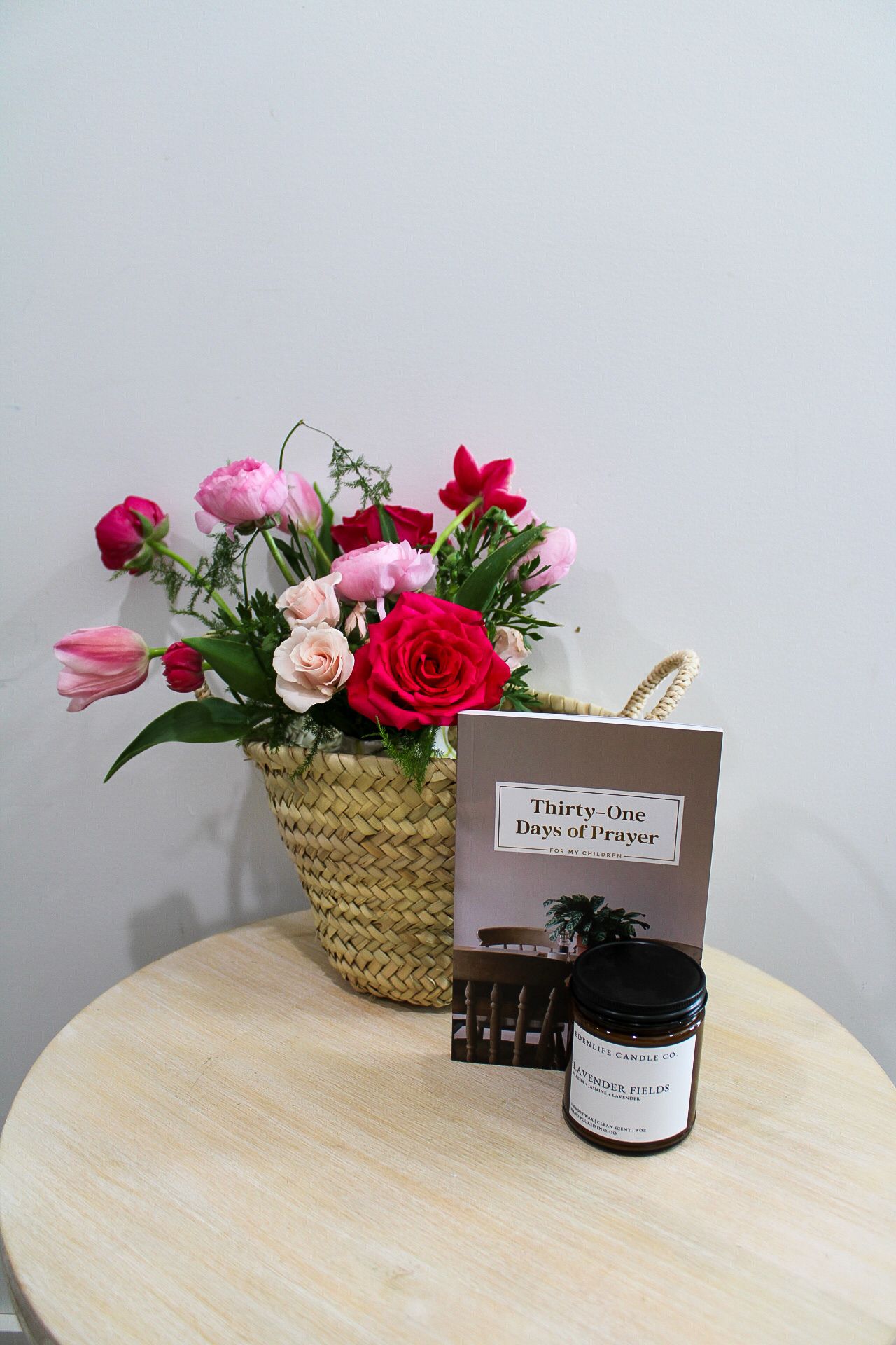 Wicker basket with pink and red roses beside a prayer book and candle on a wooden table.