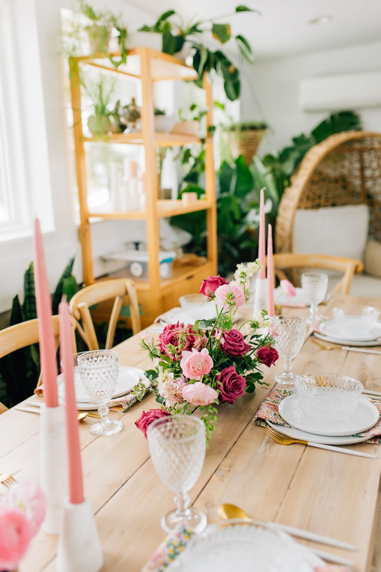 Whimsical table setting with pink floral centerpiece, taper candles, and natural wood shelving with plants.