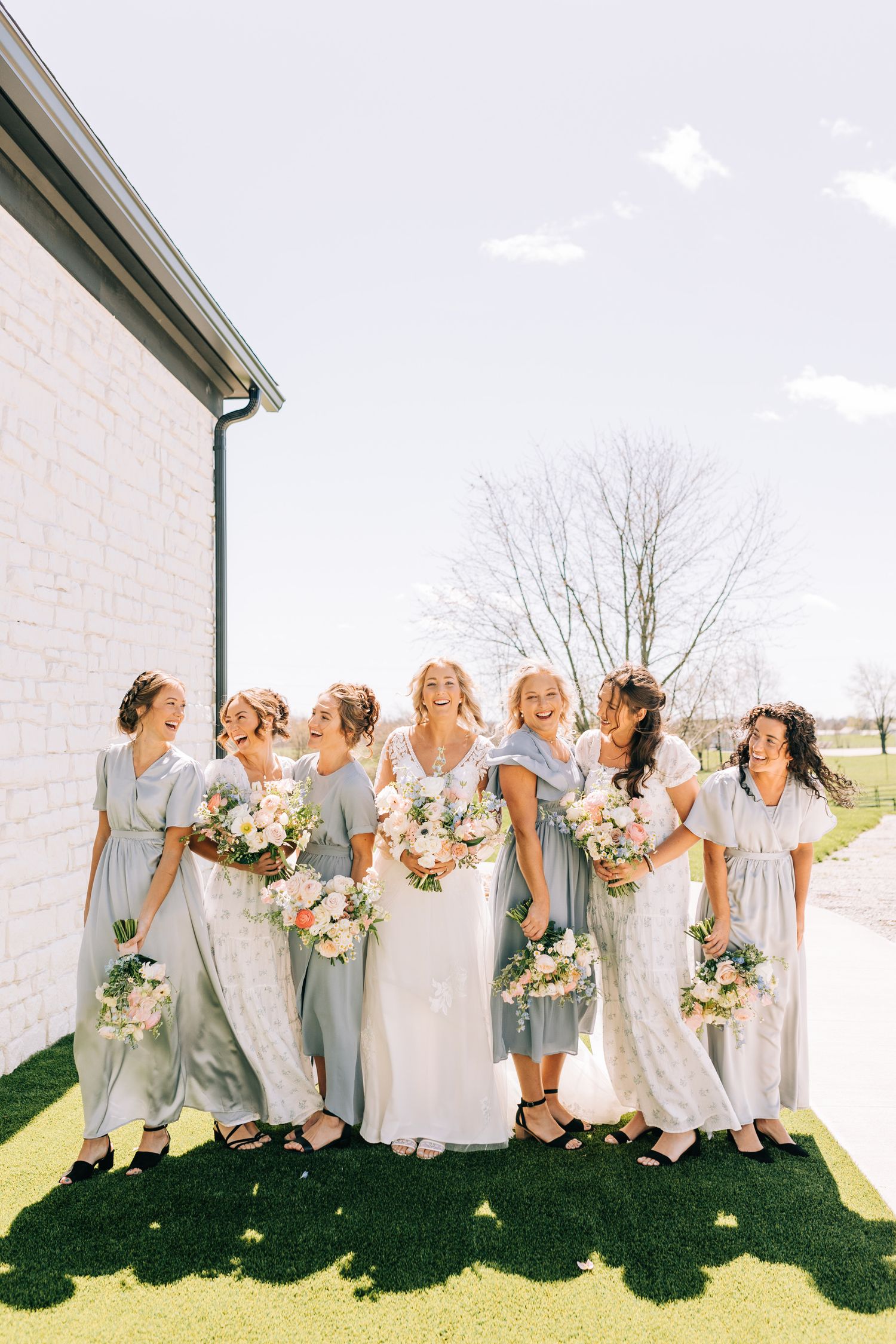 Wedding party in soft blue dresses poses together against white building in bright sunlight.