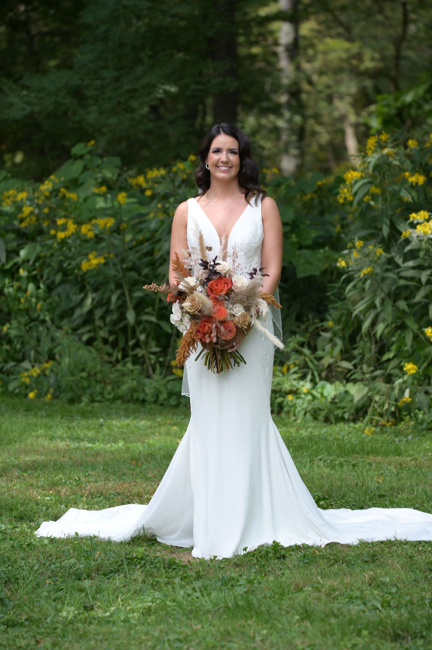 A bride in a white wedding dress poses with her autumn-themed bouquet in a garden setting with yellow wildflowers.