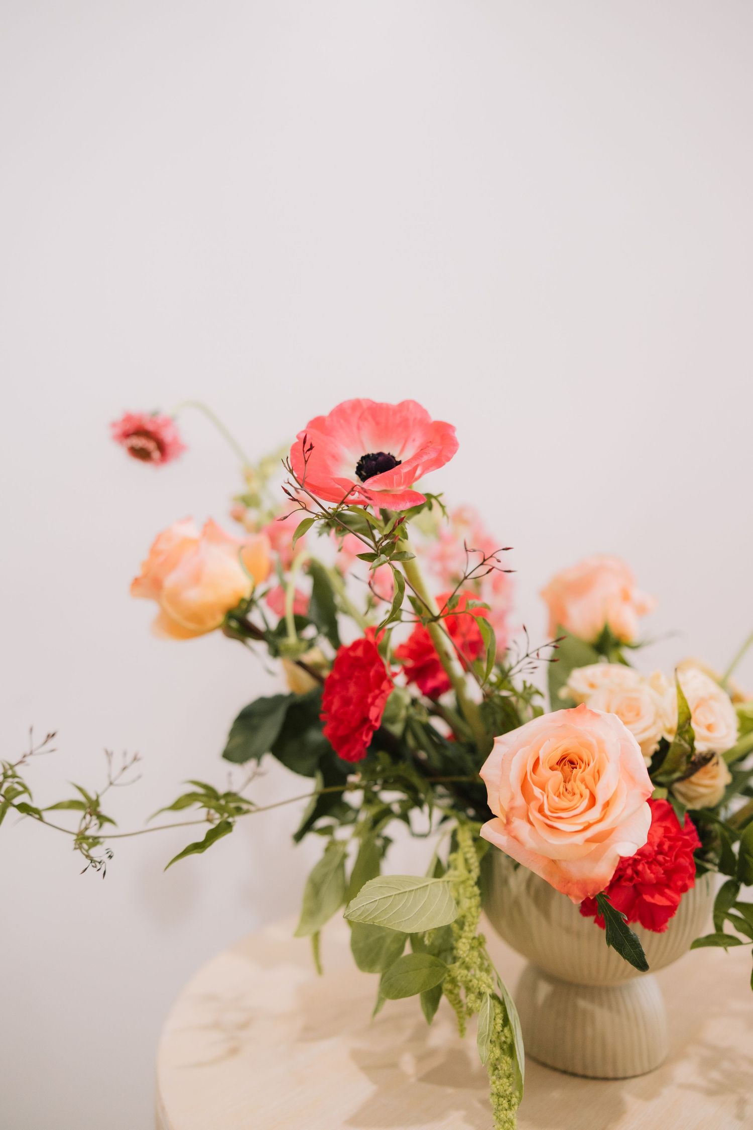 Delicate floral arrangement featuring coral roses, red carnations and greenery in white ceramic vases against light background.