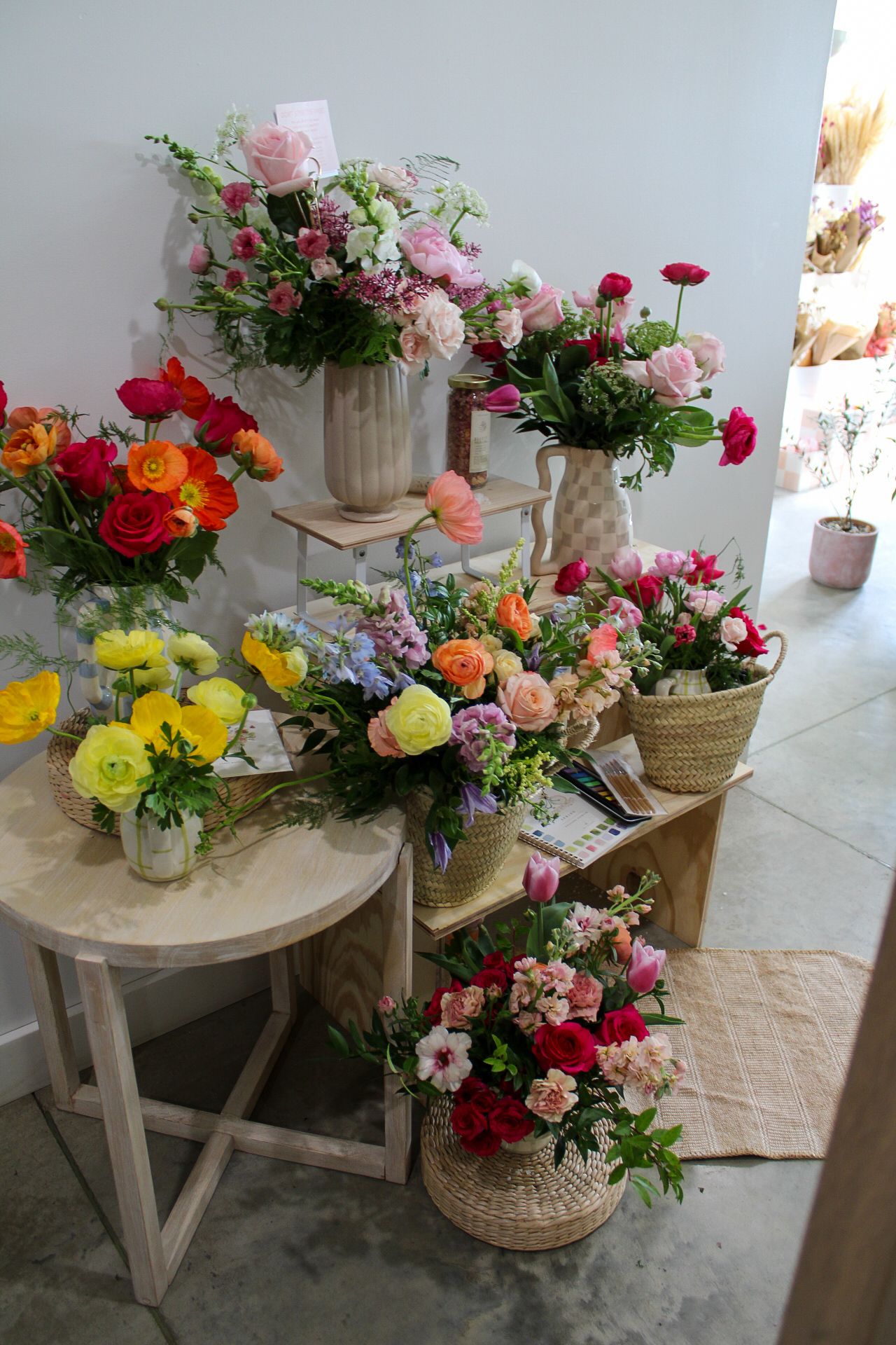 Colorful floral arrangements in vases and baskets displayed on wooden tables inside a bright flower shop.
