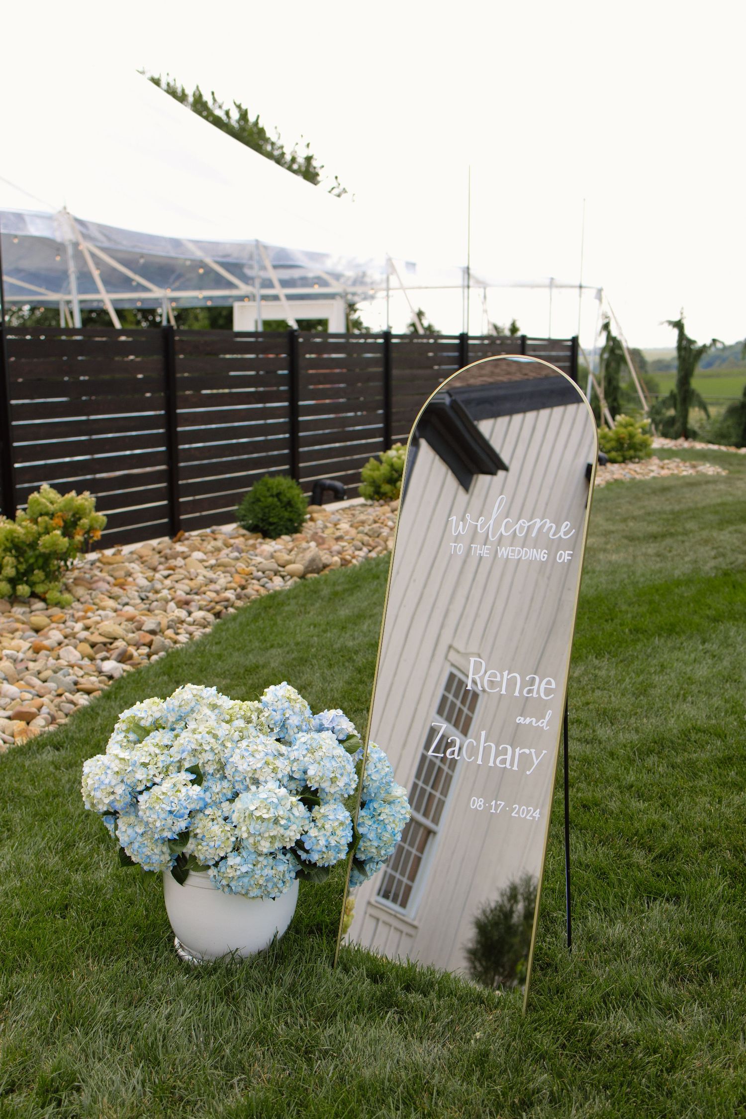 A modern wedding welcome sign with blue hydrangeas displayed on a lawn with mountain views in the background.