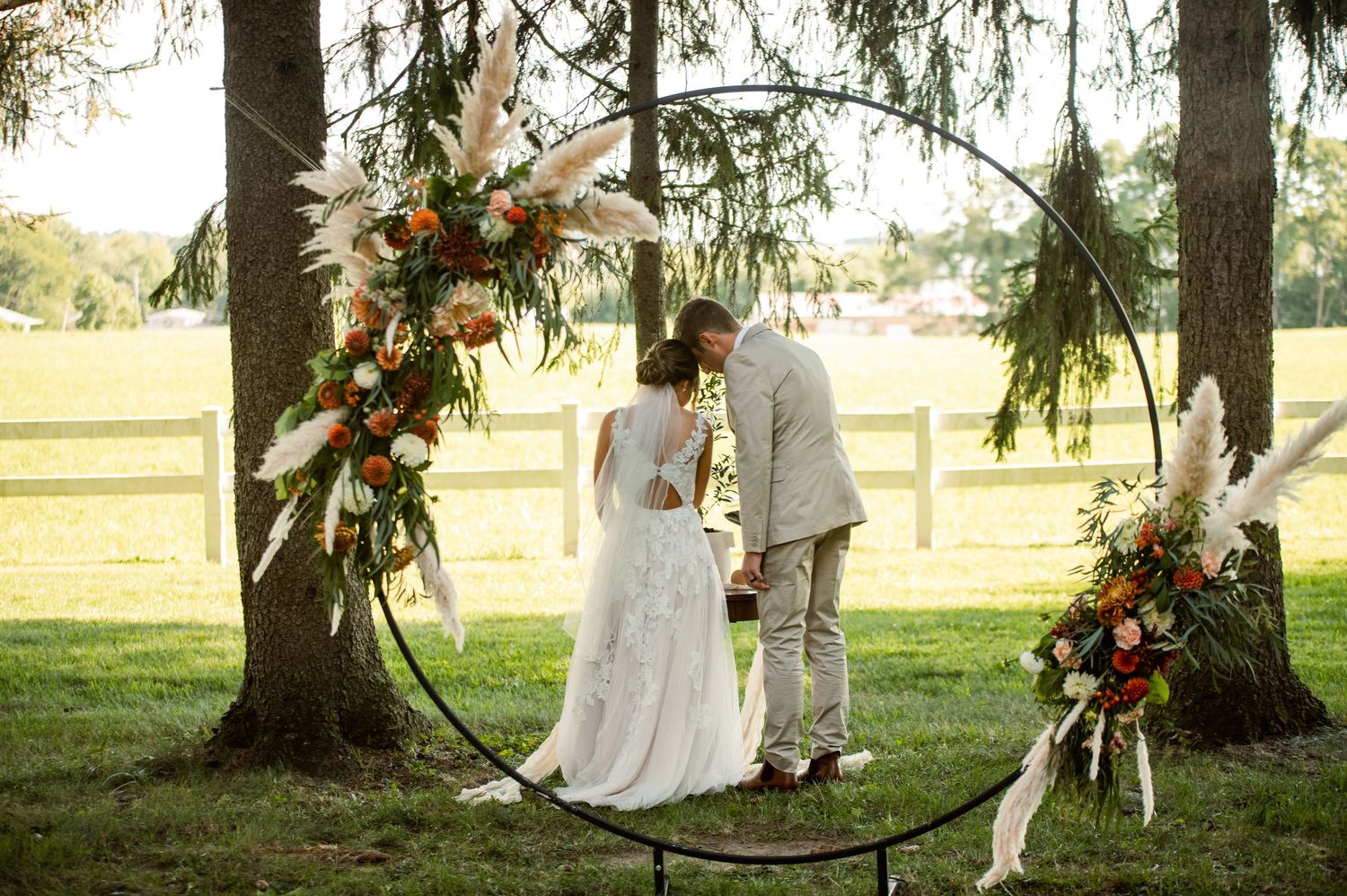 Circular metal wedding arch decorated with autumn-colored flowers in pastoral farm setting.