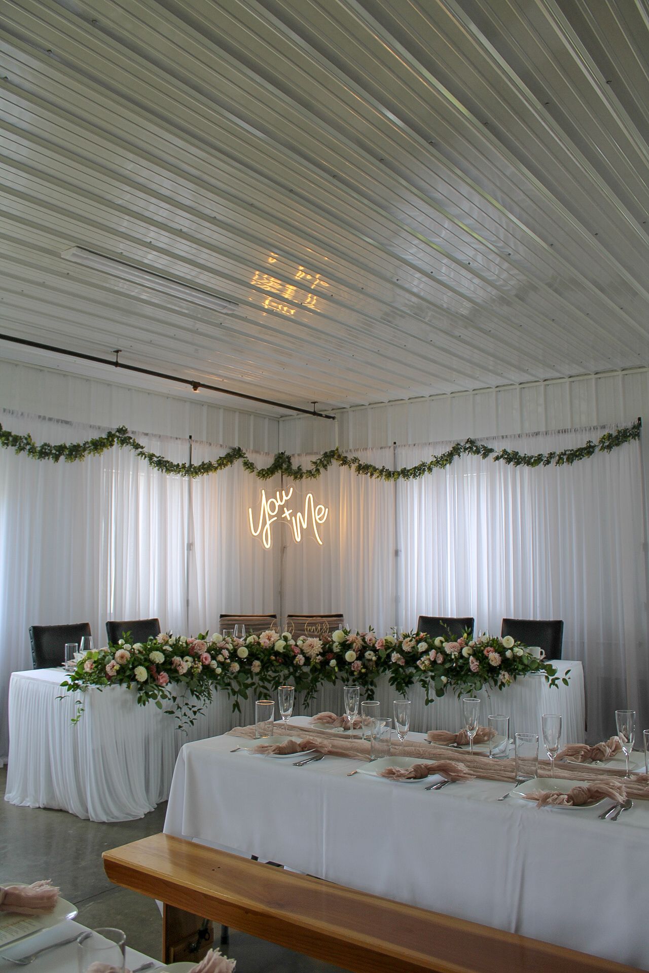 Wedding reception venue with white drapes, neon sign, and floral head table decorations.