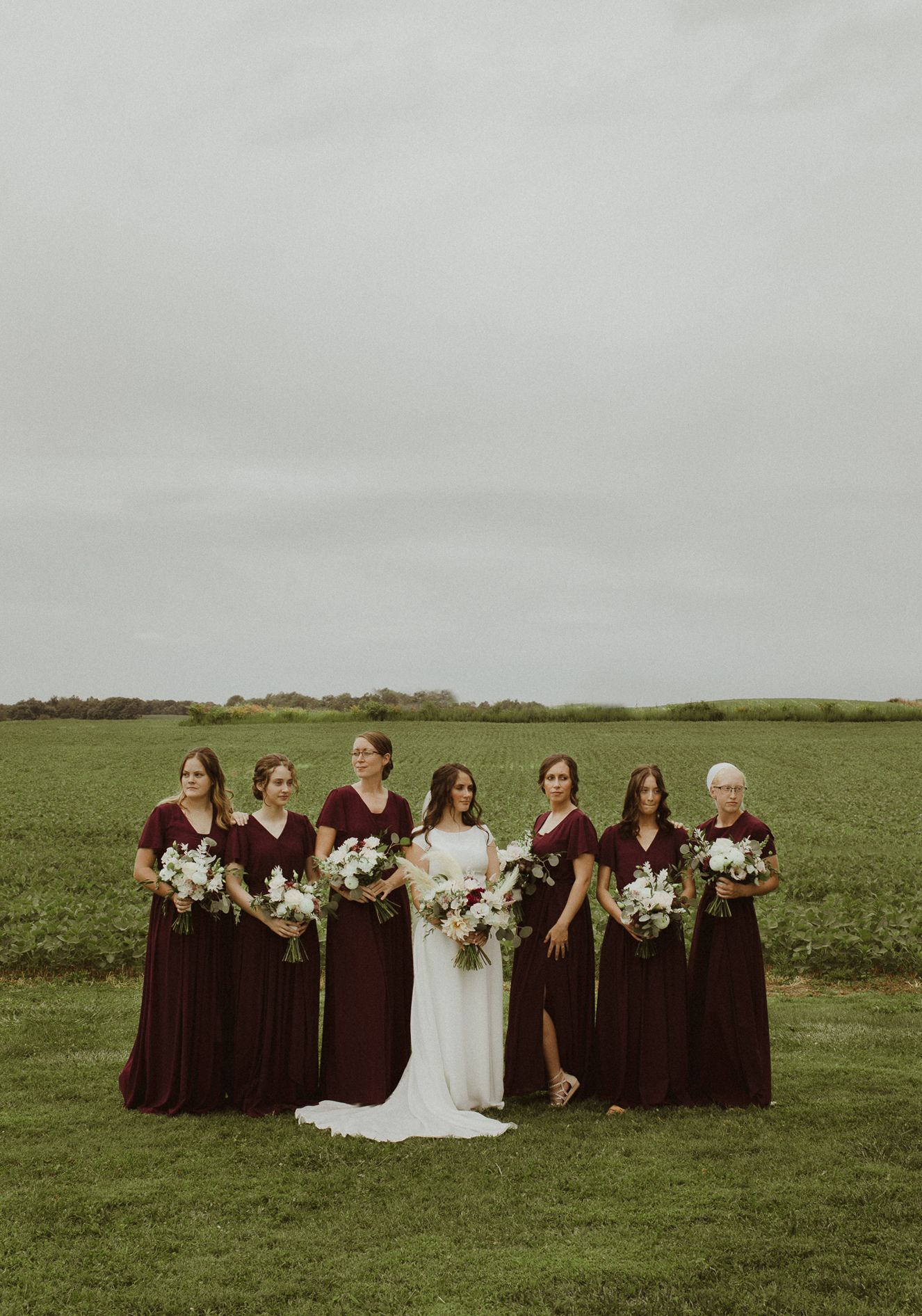 A bridal party poses in a green field with burgundy bridesmaid dresses and white floral bouquets.