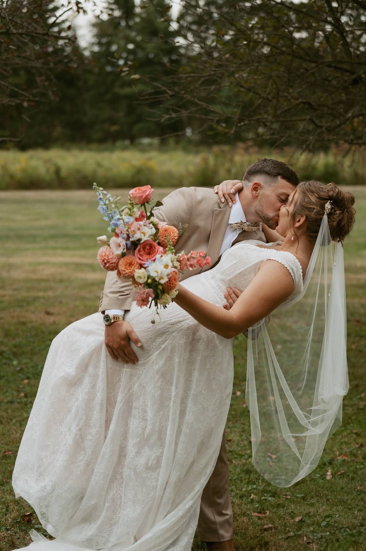 A romantic wedding couple embraces in a dip kiss outdoors, with the bride holding a coral and blue floral bouquet.