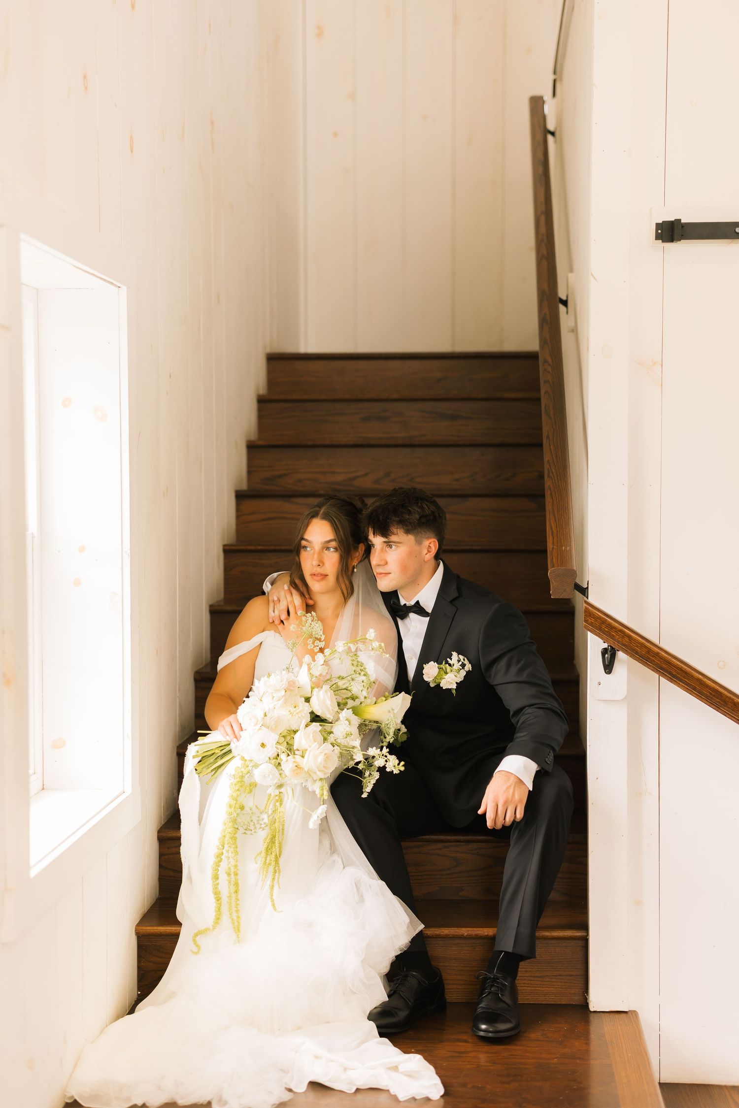 A couple in wedding attire sits together on wooden stairs in a bright, minimalist hallway with white walls.