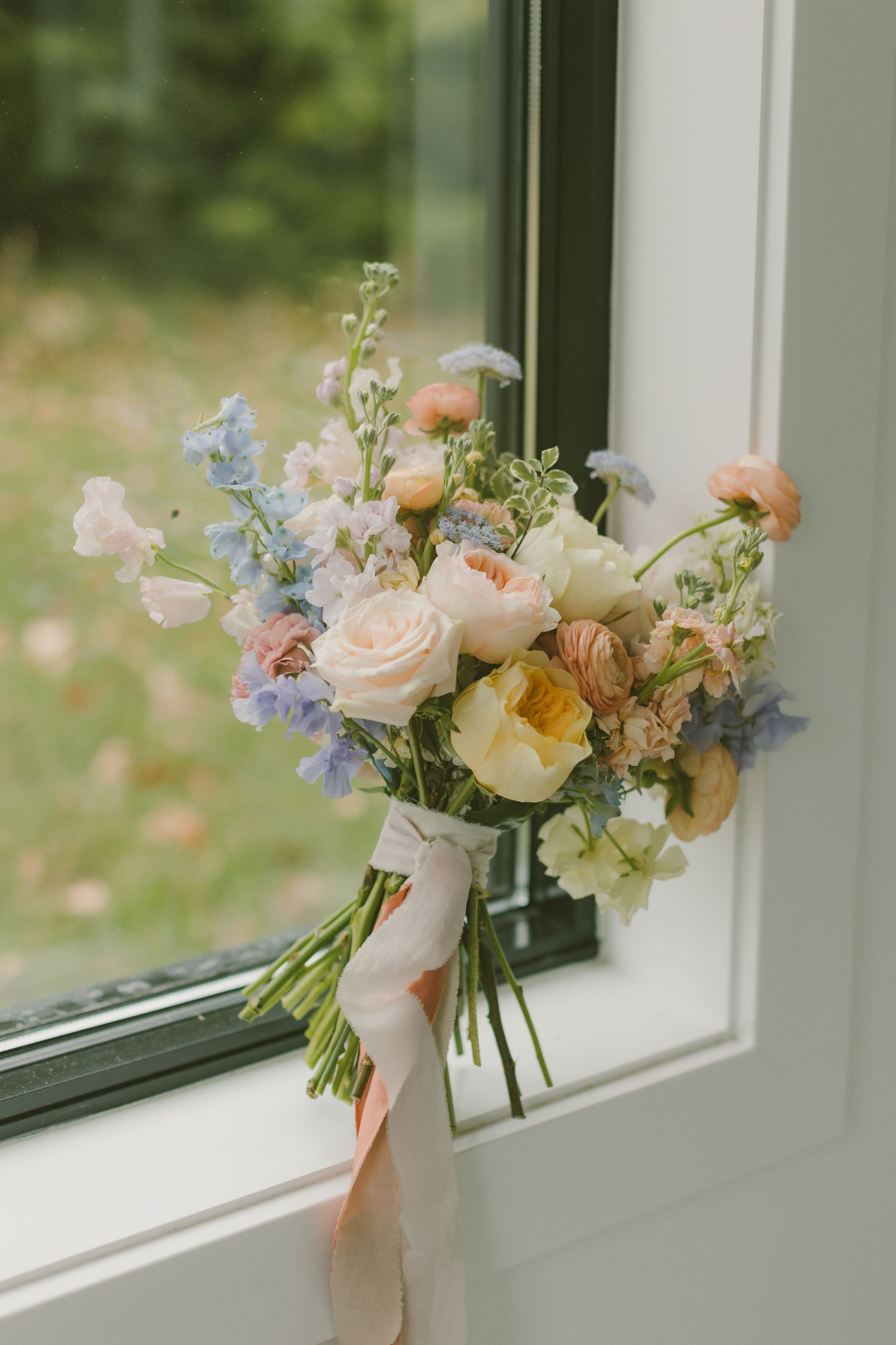 A romantic spring bouquet with peach roses and blue delphiniums tied with a silk ribbon rests on a windowsill.