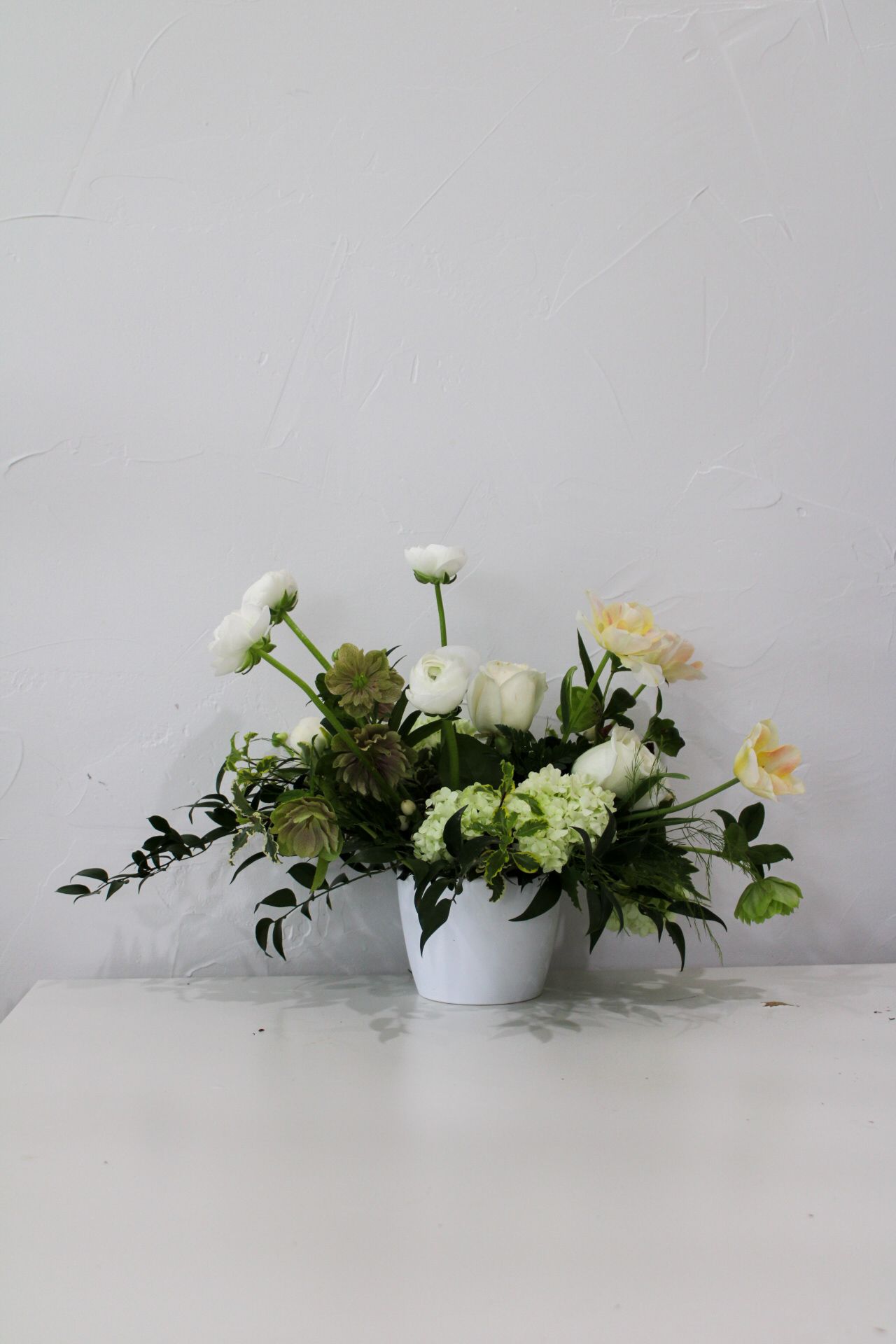Soft white and cream floral arrangement with roses and greenery in a simple white container.