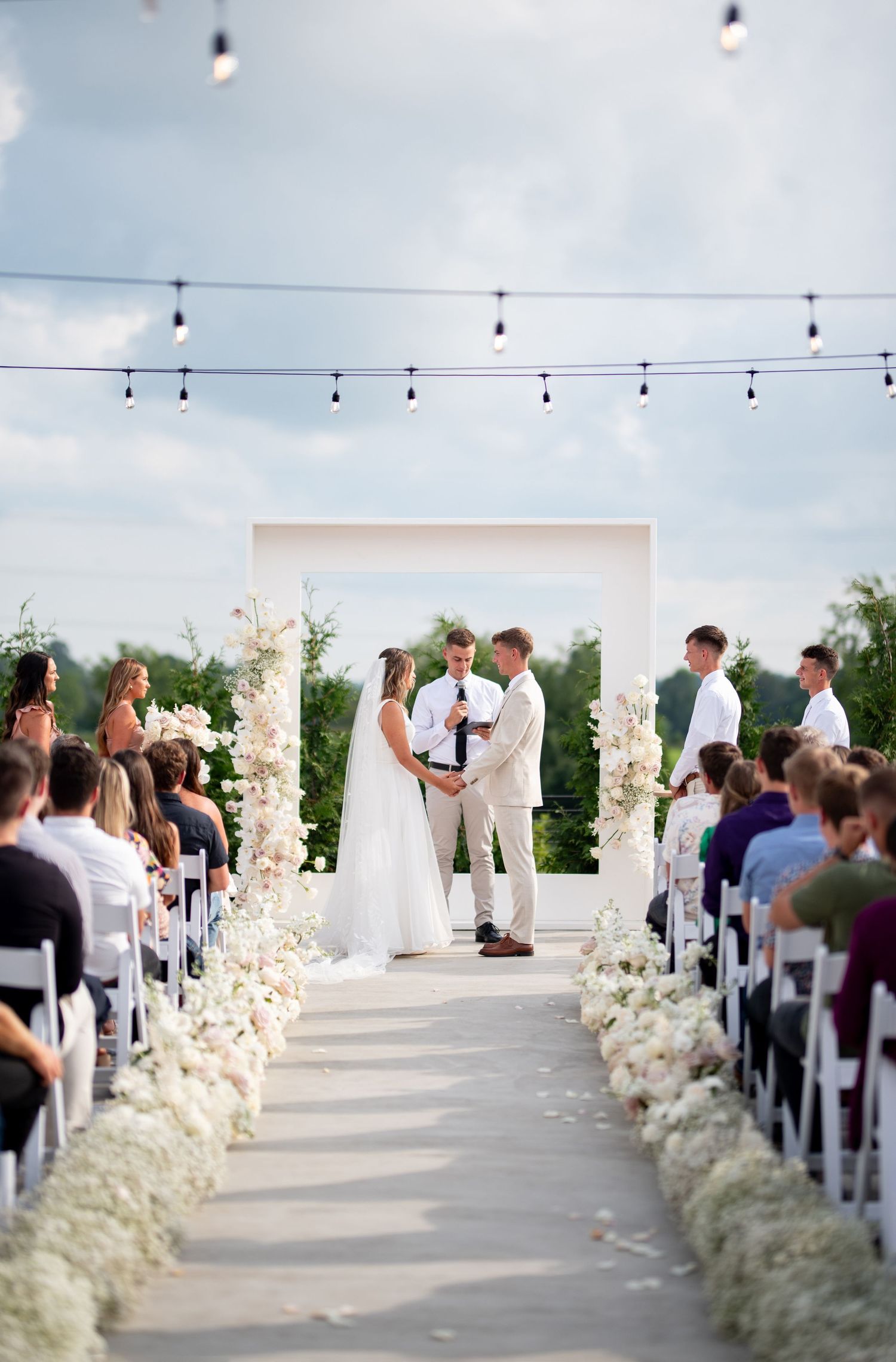 Outdoor wedding ceremony setup with string lights and floral-lined aisle under blue sky.