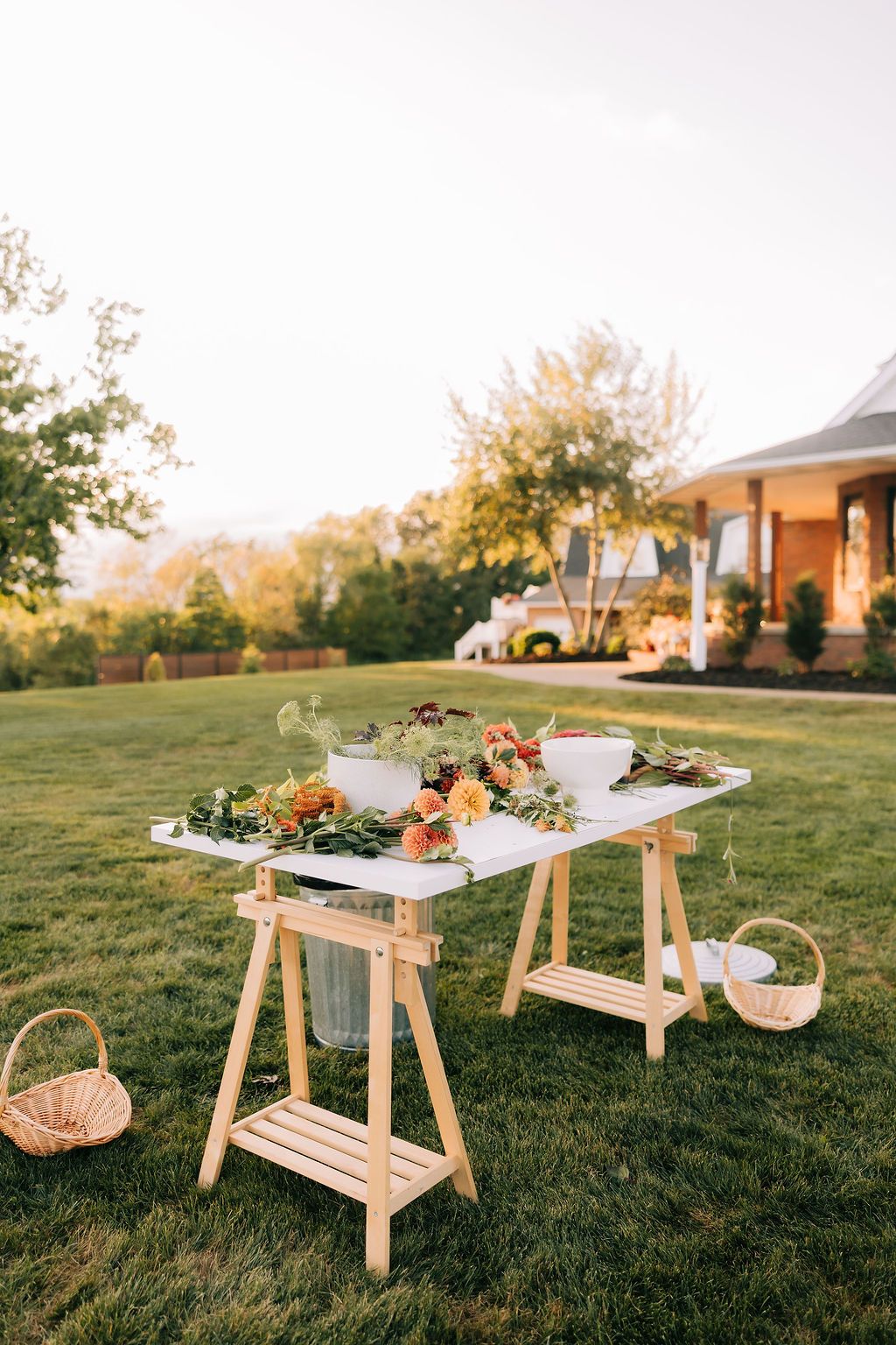 Rustic wooden sawhorse tables set up outdoors on grass with floral arrangements and place settings at sunset.