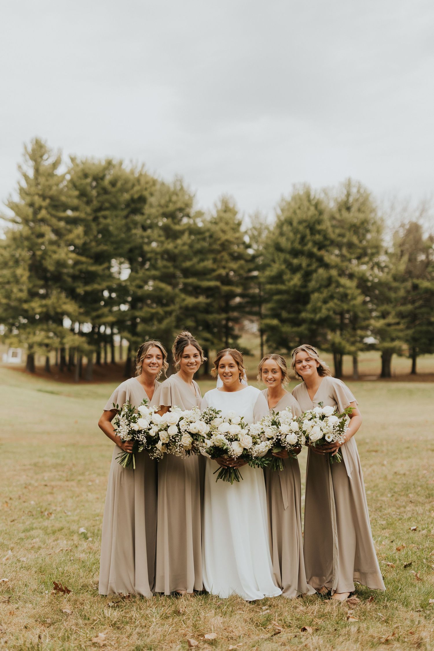 Wedding party in neutral taupe dresses stands together holding white floral bouquets in an open grassy field.