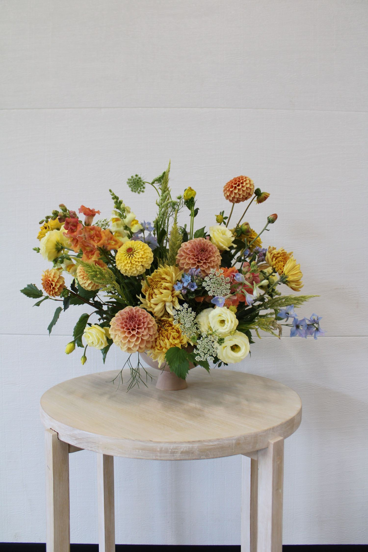 A floral arrangement of orange, yellow and pink dahlias with delicate white flowers on a light wooden round table.
