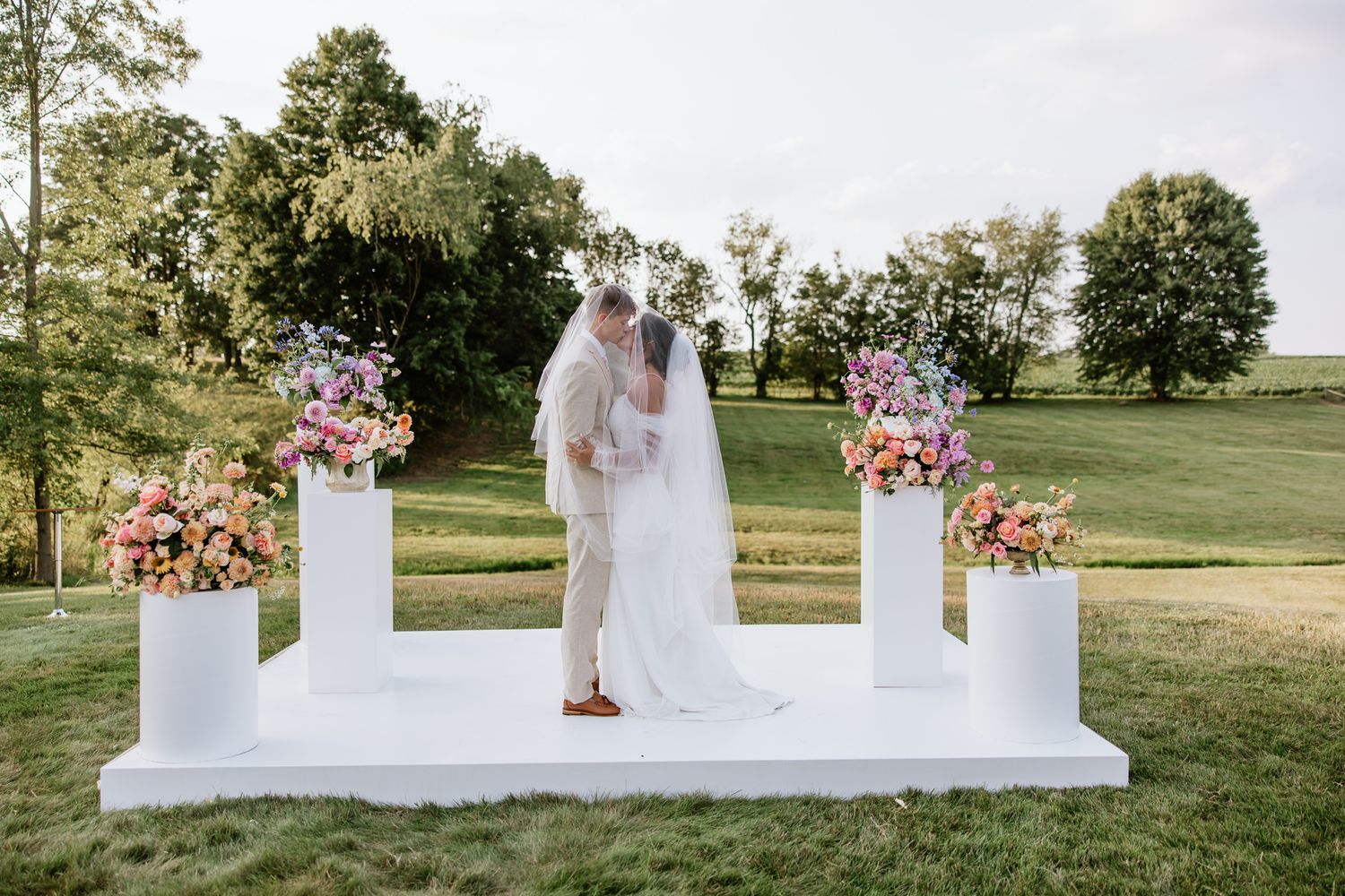 An elegant white wedding ceremony platform decorated with colorful floral arrangements in an outdoor setting.