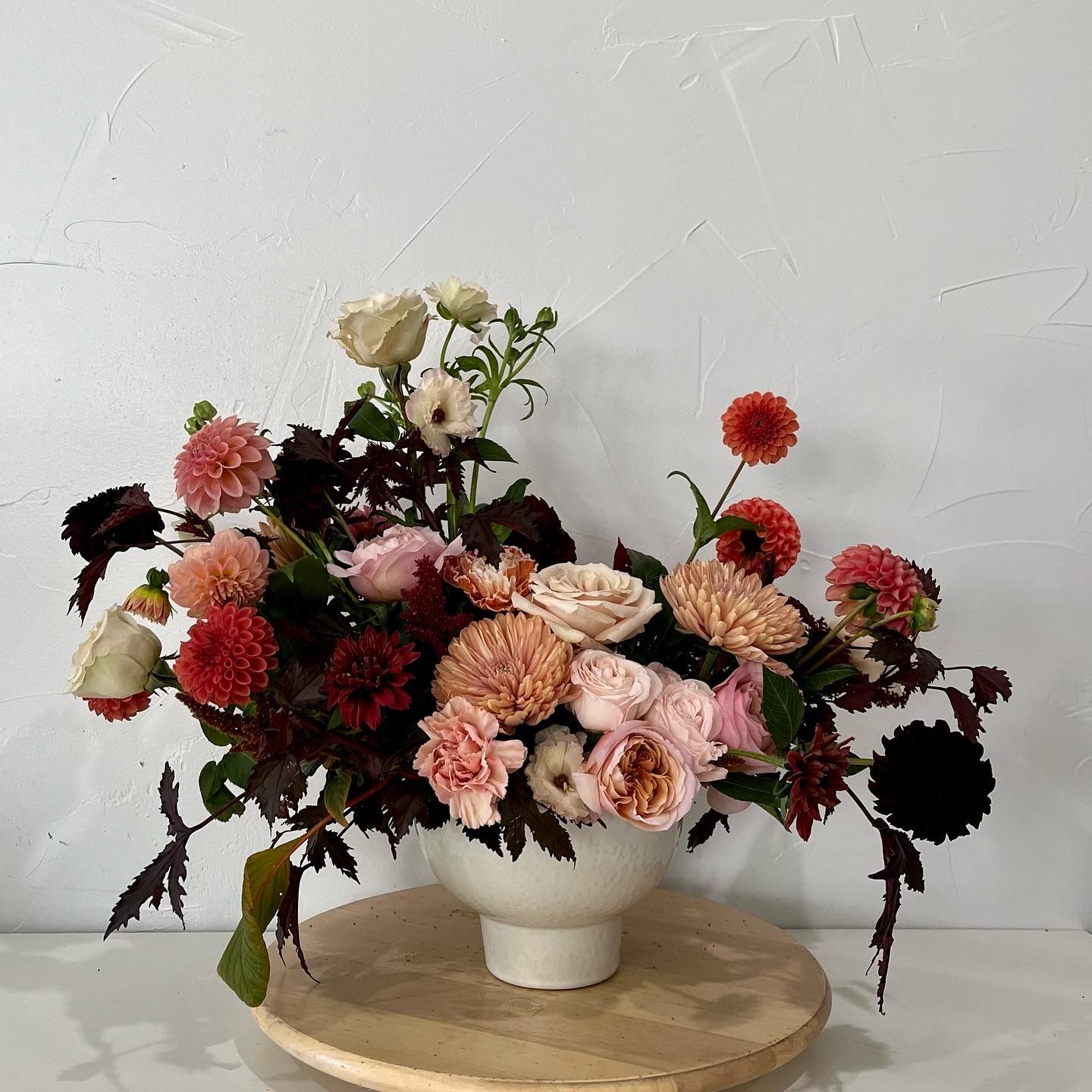 Lush arrangement of burgundy and coral flowers with dahlias in a white ceramic vessel.