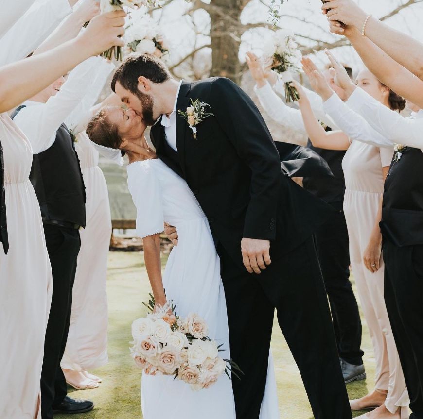 Wedding party forms archway with raised arms as newlyweds share romantic kiss underneath.
