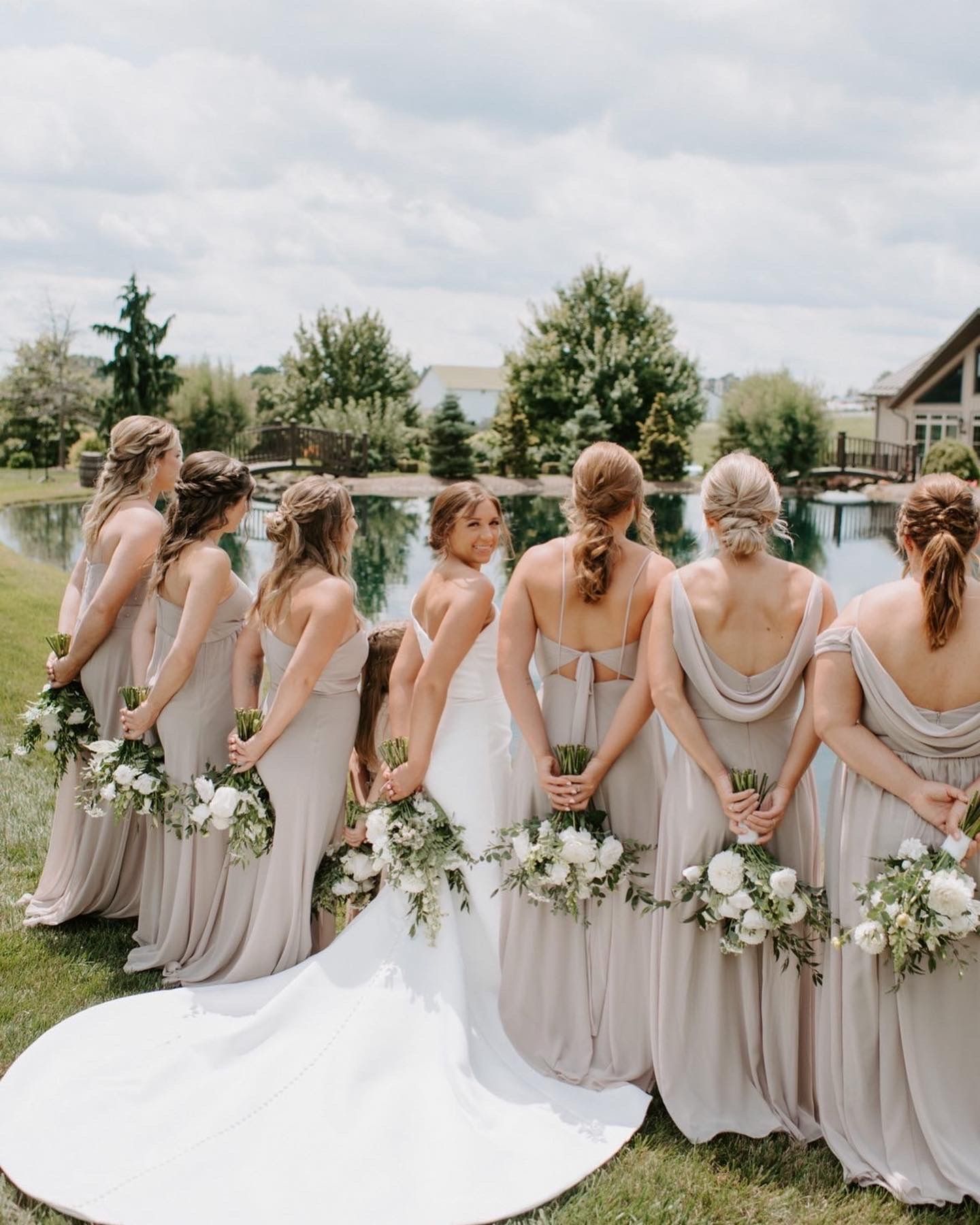 Row of bridesmaids in matching gray dresses holding white bouquets by lakeside.
