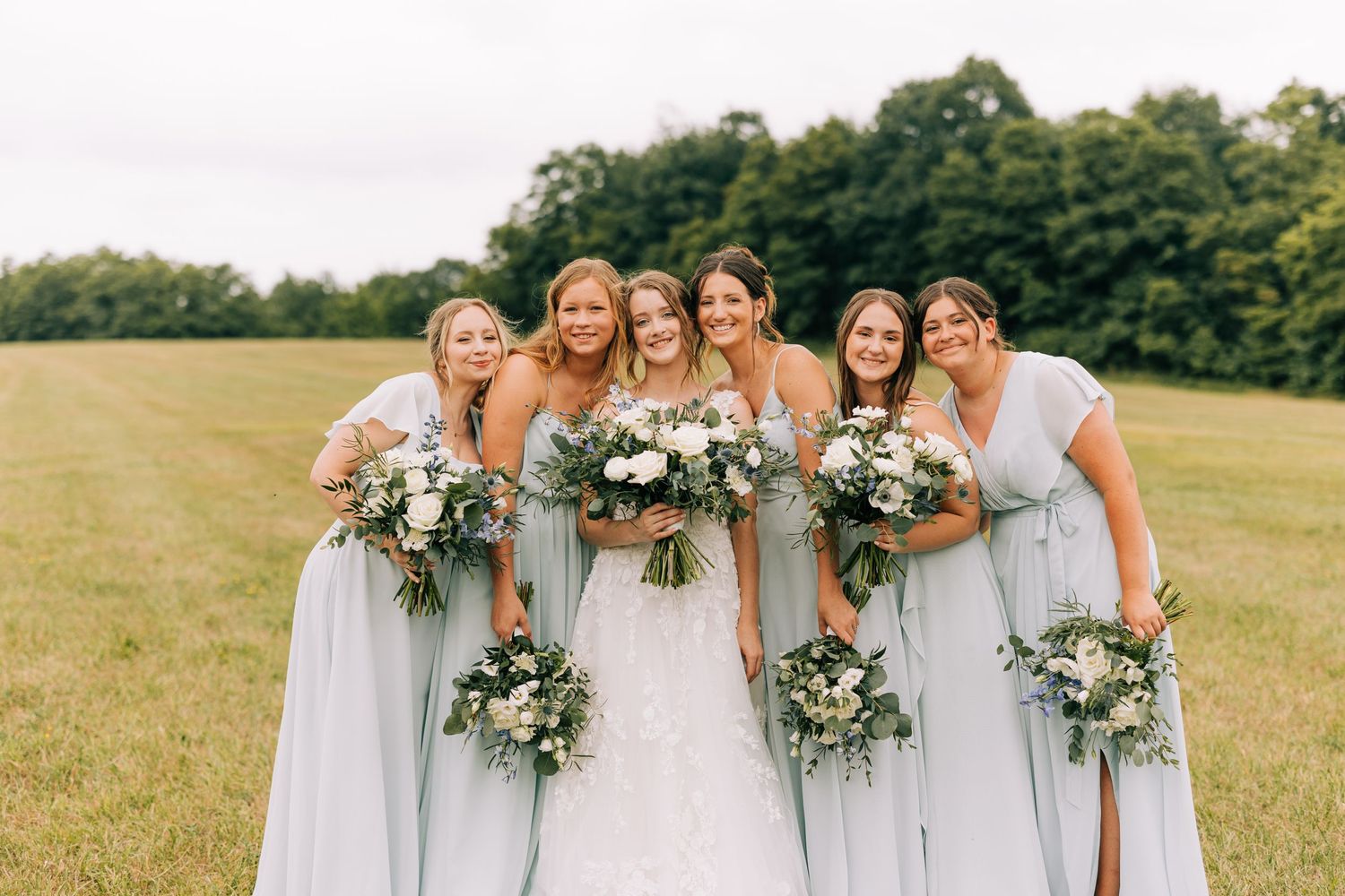 Bridesmaids in matching light blue dresses pose together holding white floral bouquets in a sunny field.