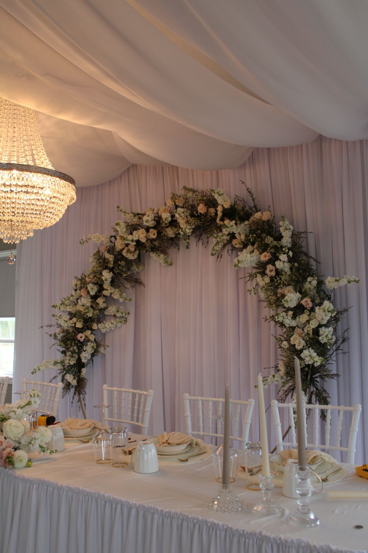 Romantic wedding reception setup with crystal chandelier and floral arch over white dining tables.