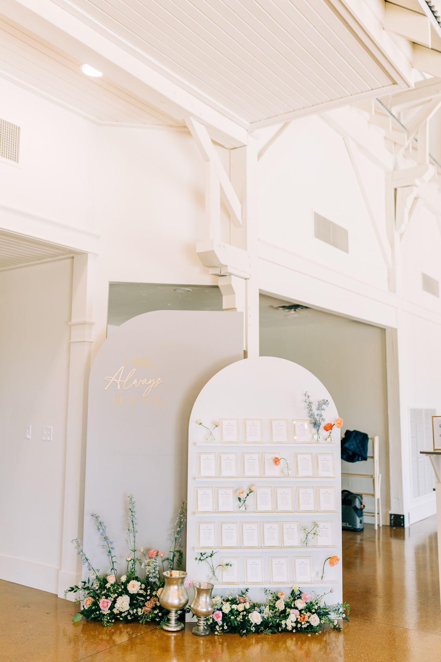 White wedding seating chart display with floral arrangements and arch detail in bright indoor venue.