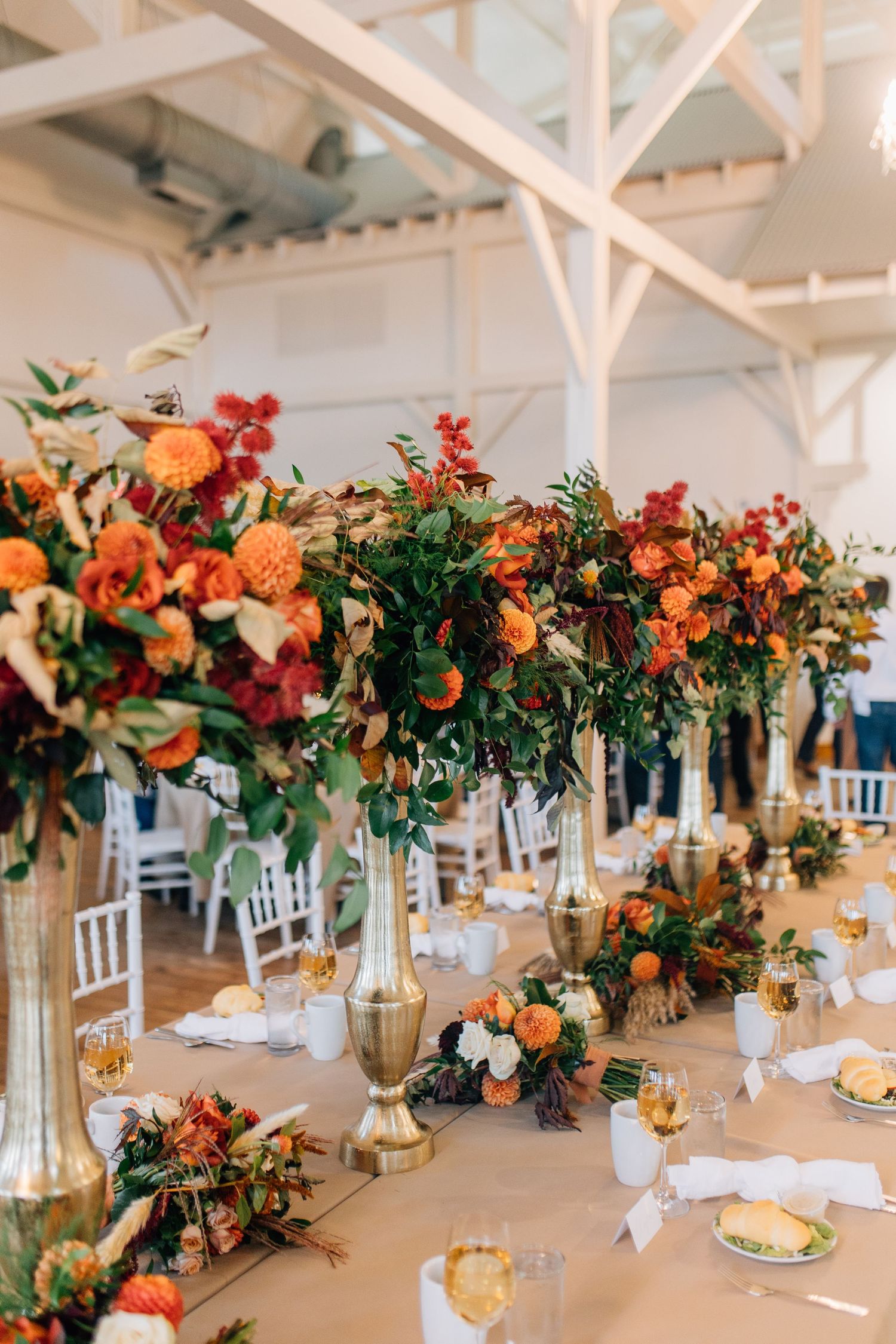 Wedding reception tables decorated with dramatic fall-colored floral arrangements in tall gold vases.