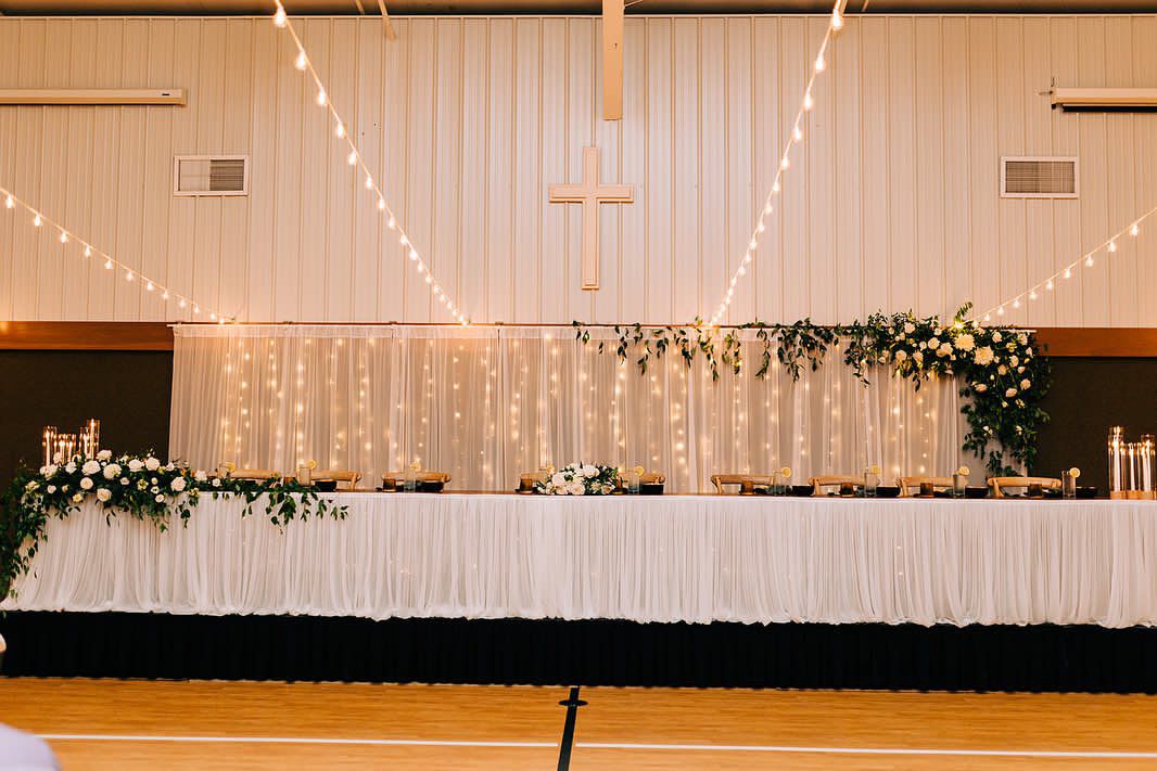 Elegant wedding head table setup with white draping, string lights and floral arrangements in a gymnasium space.