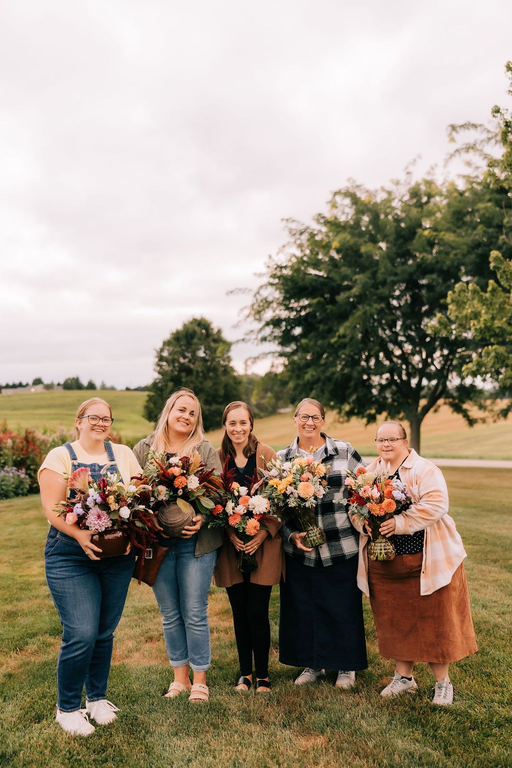 A group of bridesmaids in casual denim and brown attire holding vibrant fall-colored floral bouquets outdoors.