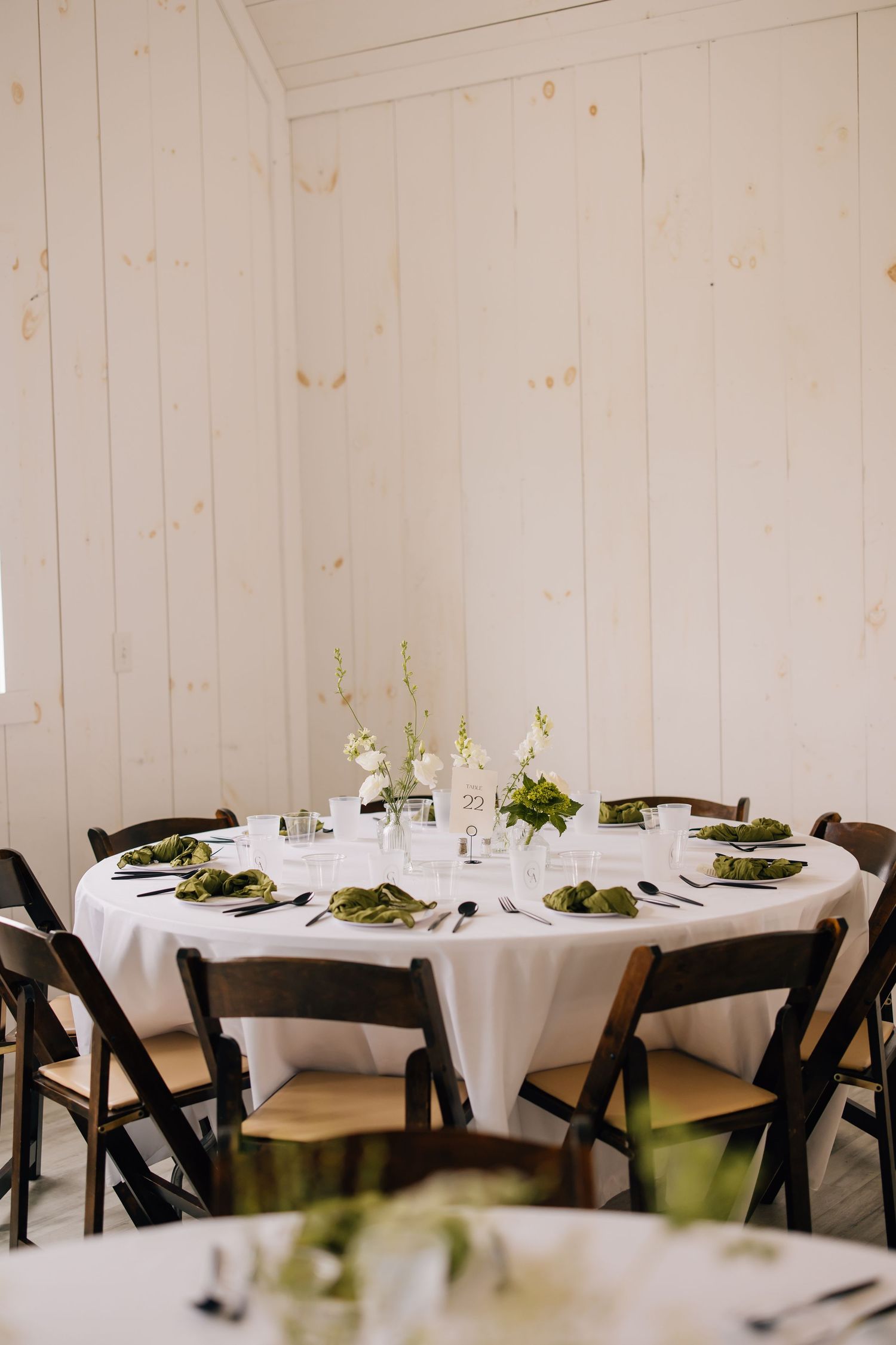 An elegant round dining table set with white linens, black folding chairs, and simple floral centerpieces in a white room.