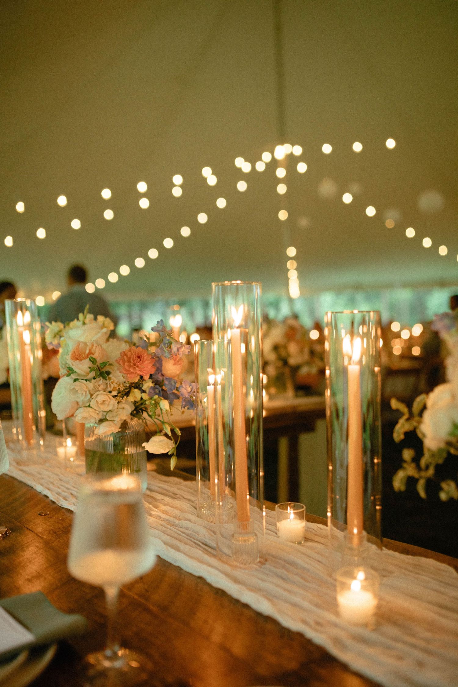 Long banquet table decorated with romantic candlelight and floral arrangements under string lights creates an elegant ambiance.