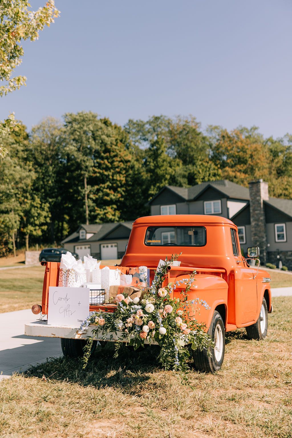 A vintage orange pickup truck sits in a grassy field with floral arrangements displayed in its truck bed.