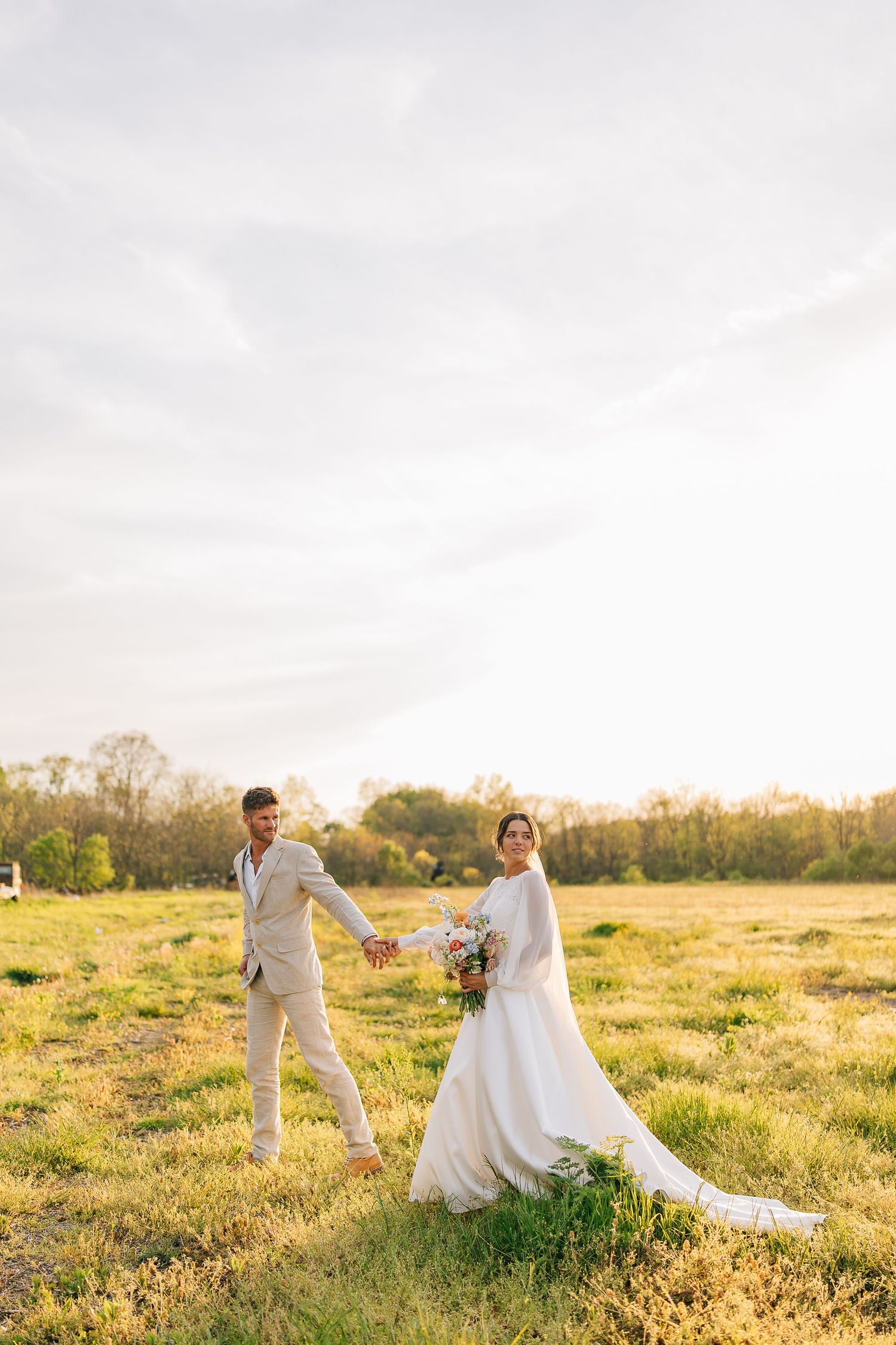 A couple in wedding attire playfully runs through a sunlit meadow at golden hour.
